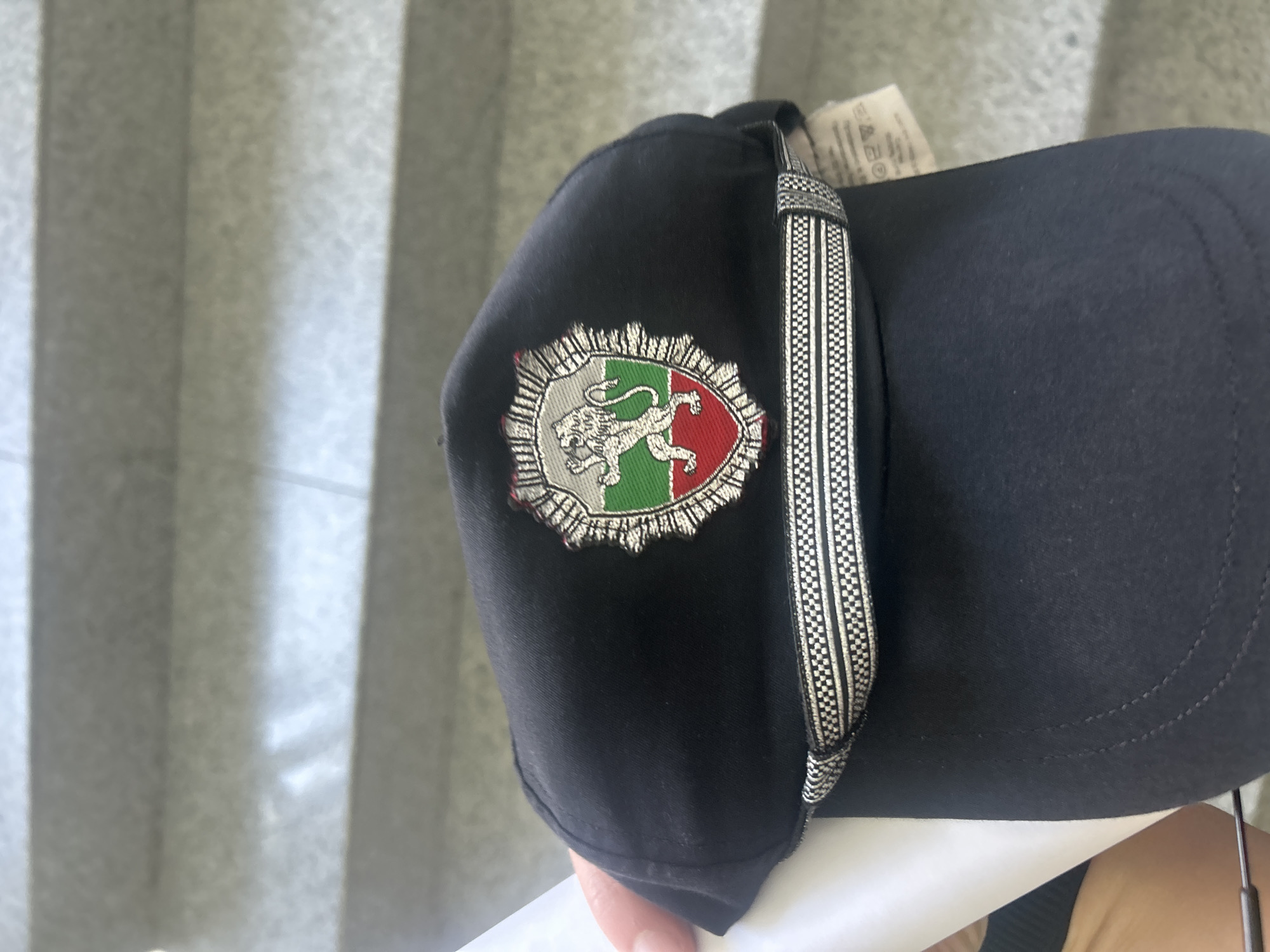 A close-up photograph depicts a black baseball cap with an embroidered patch on the front. The patch features a heraldic shield design, containing a white lion rampant over a green field, adjacent to a red field, enclosed within a silver filigree border. A decorative strap with a black and white checkered pattern is attached to the cap, extending vertically. The background consists of a blurred gray curtain with vertical pleats. A hand holding the cap is partially visible at the bottom of the frame. The object appears to be a souvenir or novelty item featuring the Bulgarian coat of arms. There is no discernible evidence to pinpoint the location as the city of Sofia, Bulgaria.
