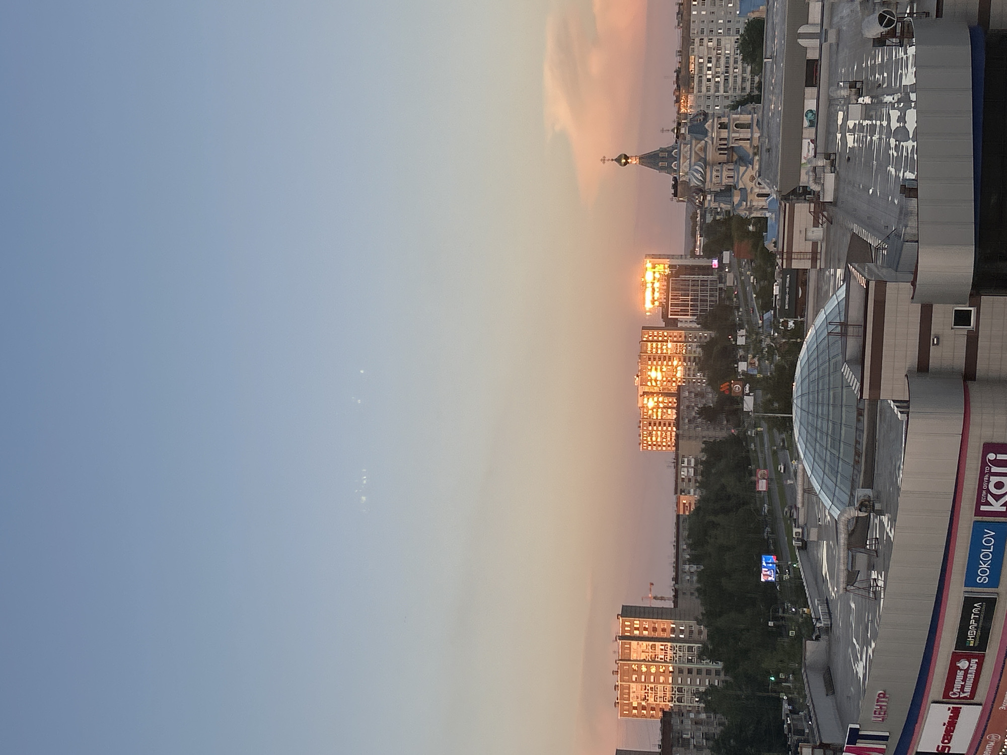 The image depicts a cityscape at sunset in Izhevsk, Russia. In the foreground, the rooftop of a shopping mall with a distinctive glass dome is visible. Various signage is present on the mall's exterior, including text in Russian such as "ЦЕНТР" (CENTER), "Sokolov", and "Kari". In the middle ground, a busy street with traffic can be seen, flanked by trees and residential buildings. The setting sun reflects brightly off the windows of several tall apartment buildings, creating a striking visual effect. To the right of the apartment buildings, a blue-domed church with golden crosses stands prominently against the twilight sky. The sky itself transitions from a clear pale blue at the top to warmer hues of orange and pink near the horizon, with subtle clouds scattered across. The overall scene captures an urban environment bathed in the warm light of dusk.