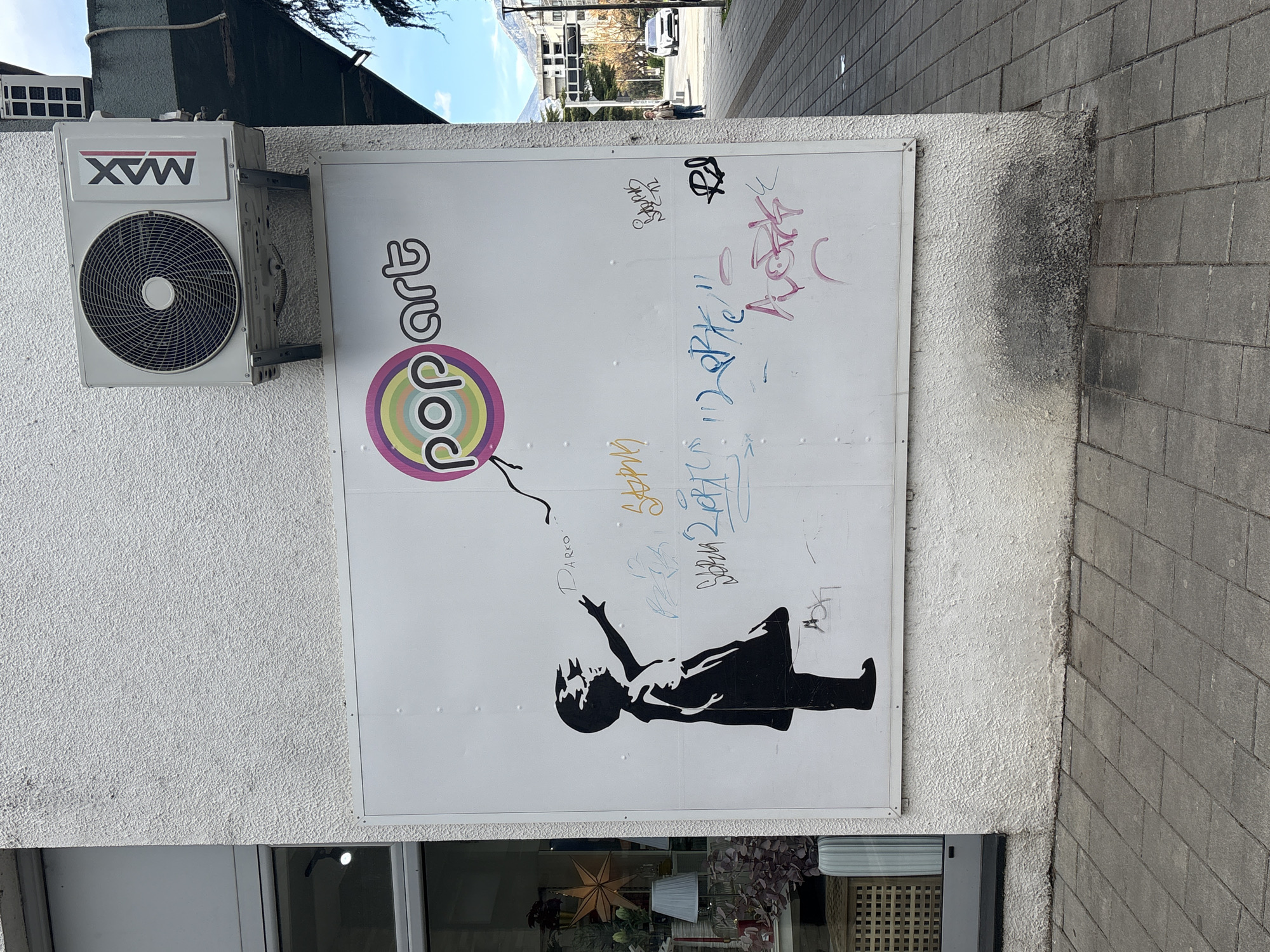 A medium shot depicts a white wall with a commercial sign on a building exterior. The sign features the word "pop art" in stylized colorful letters above a monochrome stencil of a girl reaching for a balloon. Graffiti is present on the sign's surface. To the left, an air conditioning unit is mounted on the wall. Below the sign, a display window reveals decorations inside the building. The sidewalk and street scene beyond are blurred, suggesting an urban setting, specifically Bar, Montenegro. The light conditions appear to be daylight with a clear sky visible above.