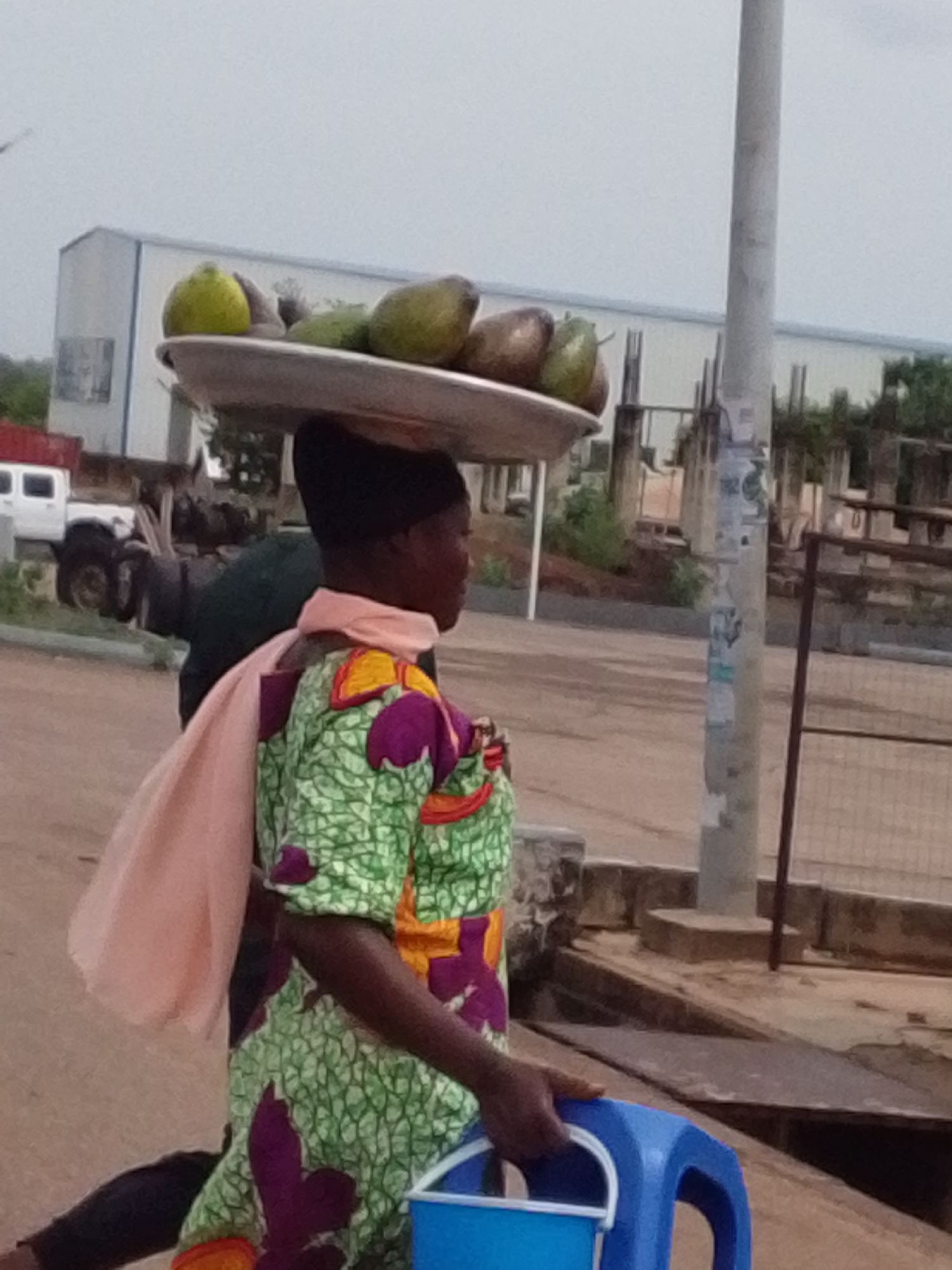 A woman carries a tray of fruit on her head and a blue plastic bucket in her hand. She wears a colorful dress with a green and purple pattern, and a light pink scarf. The background includes a large white building, a utility pole, and a vehicle. The location is Dungu, Ghana.