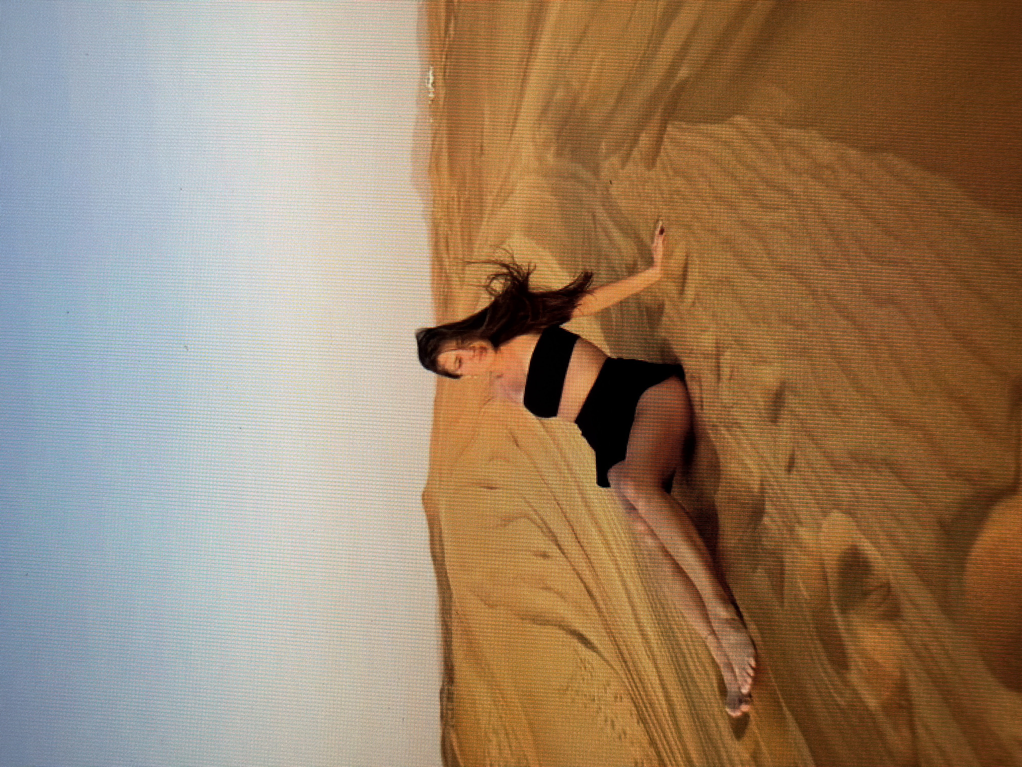 A woman with long, dark hair is sitting on a sand dune, wearing a black bikini. Her body is angled away from the camera, with her legs stretched out in front of her. The sand dune is golden brown, with shadows creating ripples and texture. The sky above is a pale blue, transitioning to a lighter shade near the horizon. In the distance, more sand dunes are visible, with faint outlines of hills or mountains. The overall scene is one of a desert landscape.
