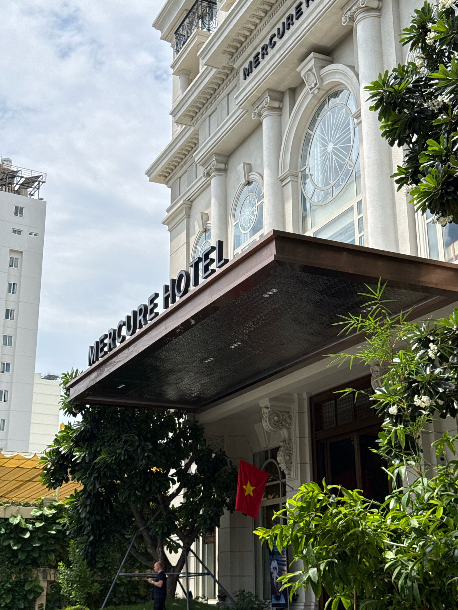 The image captures the exterior of the Mercure Hotel in Nha Trang, Vietnam. The hotel features a white, classical-inspired facade with large, arched windows and decorative columns. A prominent sign above the entrance reads "MERCURE HOTEL" in large, black lettering. A dark-colored awning extends over the entrance. Lush green trees and foliage frame the hotel, with some trees adorned with small white flowers. A Vietnamese flag, red with a yellow star, is visible hanging from the building to the left of the entrance. In the foreground, a person dressed in dark clothing is standing, holding a phone. To the left of the hotel, a tall, modern white building with multiple windows is partially visible against a cloudy sky. The overall scene suggests a well-maintained hotel entrance in a city environment, likely during daytime.