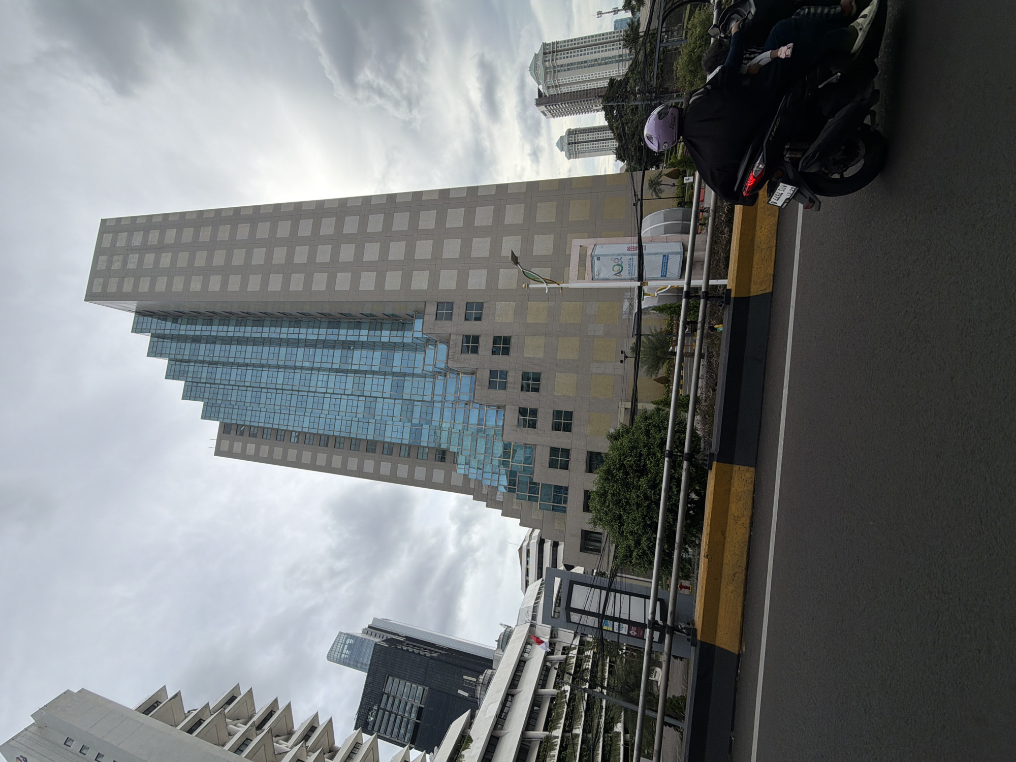 The image, taken in Central Jakarta, Indonesia, captures a street view dominated by tall buildings under an overcast sky. On the right side of the frame, a motorcycle with two riders is visible on the road. The motorcycle has the license plate "D 4462 SUM". A yellow and black barrier separates the road from the sidewalk. In the background, a prominent building with a tiered, glass facade stands out. Several other tall buildings are visible, including one with a patterned exterior. Overhead, utility lines cross the scene.