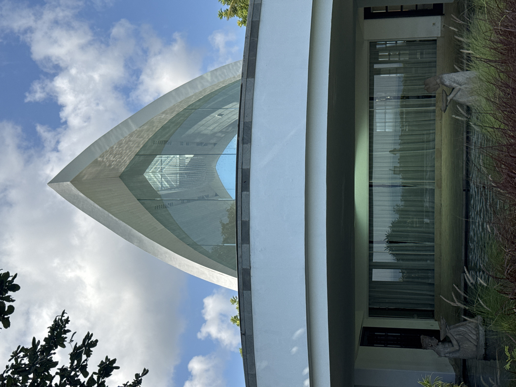 The image features the architectural marvel of the "The Ritz-Carlton, Bali, Chapel", located in Kuta Selatan, Indonesia. The chapel's design is characterized by a sharp, triangular structure primarily composed of glass and a reflective gray material. This structure rises against a backdrop of a partly cloudy sky. Adjacent to the chapel is a multi-level building with white walls and large glass windows. In the foreground are two stone statues set amidst greenery, with pampas grass prominent. The overall scene appears modern, serene, and architecturally significant.
