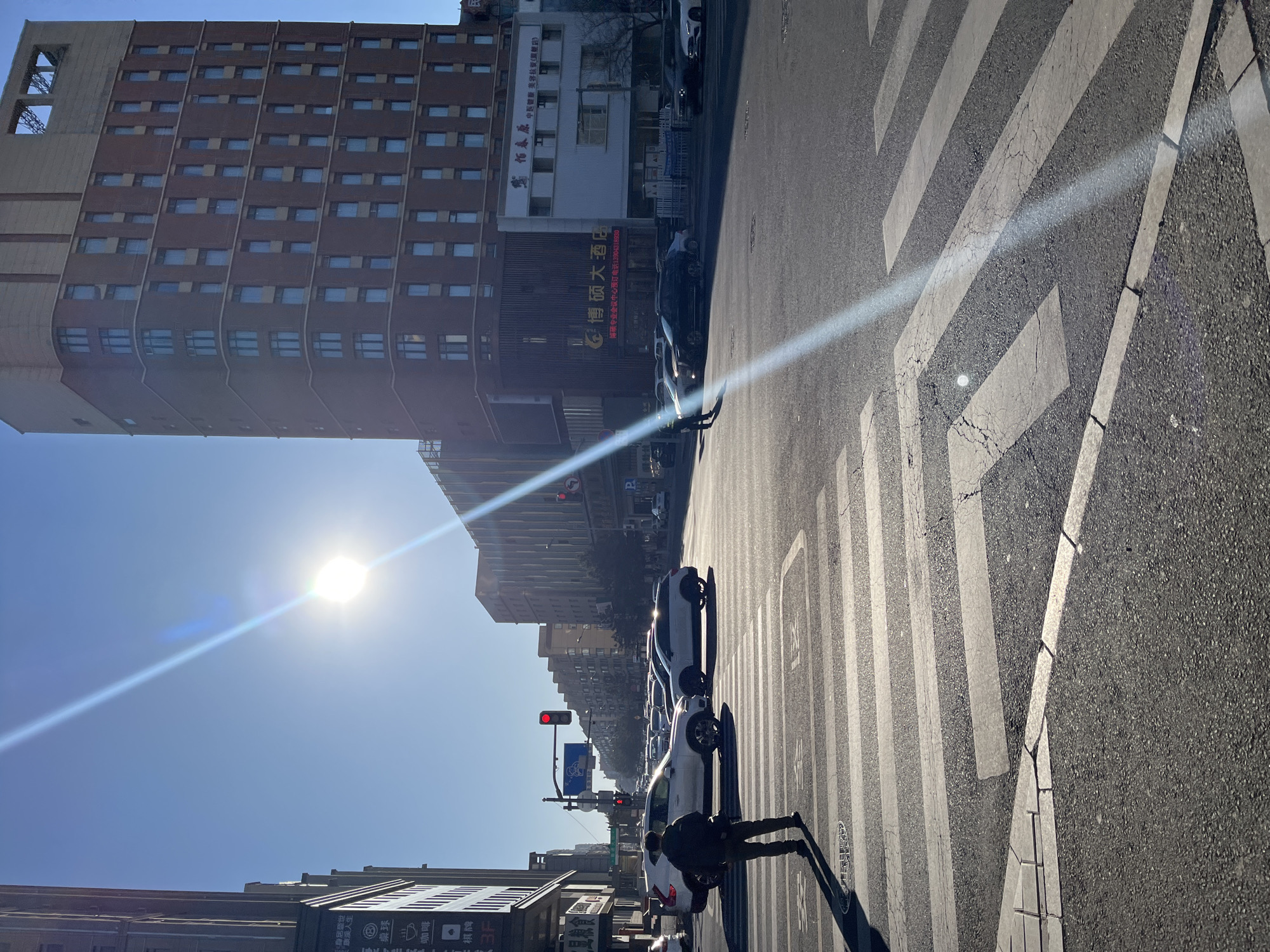 The image shows a street scene in Changchun City, China, with buildings, a crosswalk, cars, and a person crossing the street. The sun is shining brightly, creating a lens flare across the image.
On the left side of the frame is a tall building with multiple windows. A building with signs that read "博硕大酒店" and "怕春原中医" is situated next to it.
Several white cars are lined up on the street, and a pedestrian dressed in dark clothing is walking across the crosswalk. A street light with a red light can be seen down the road.
The image is taken from a low angle, looking up towards the buildings and the sky.