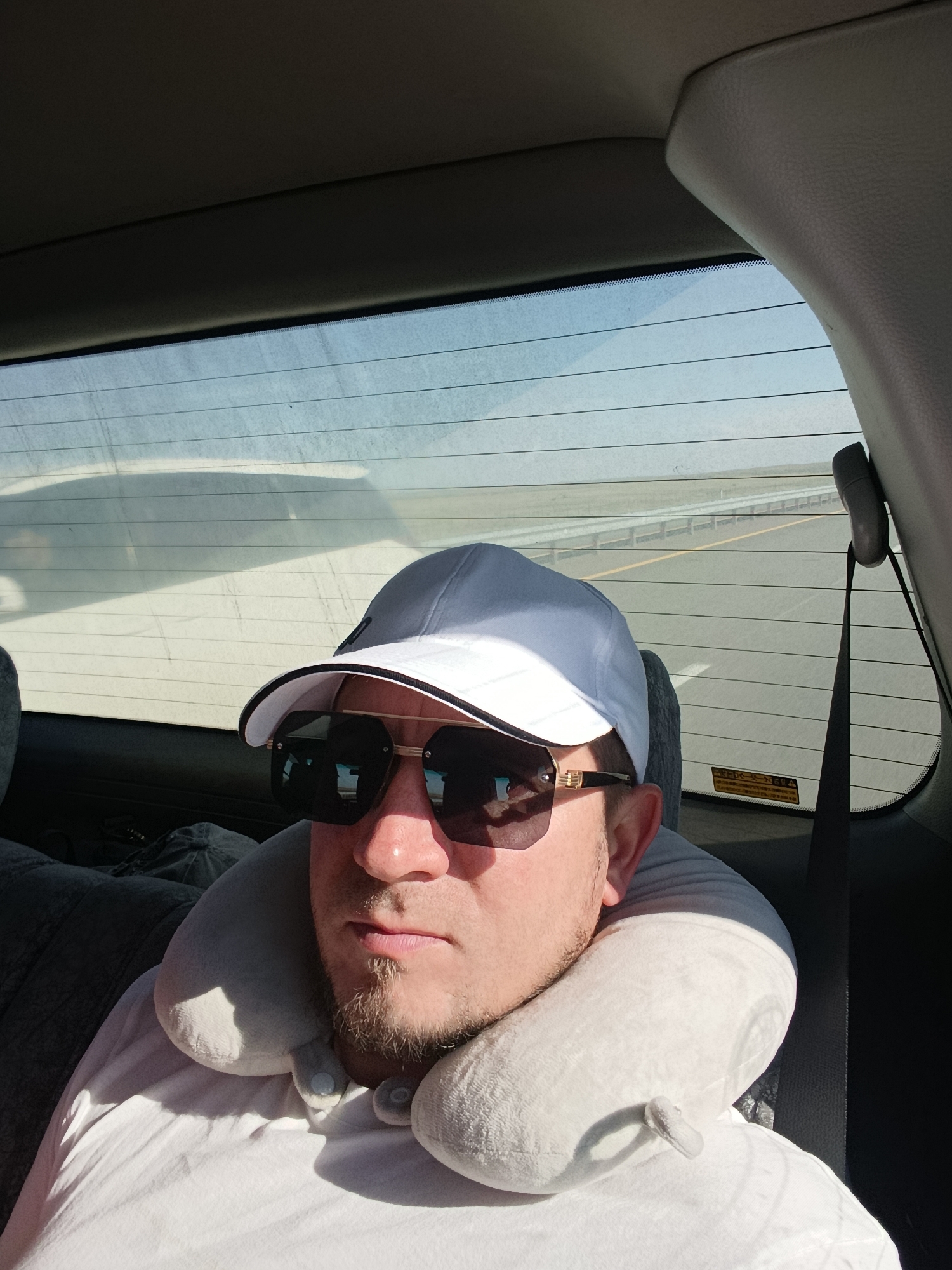 A man wearing a white baseball cap and sunglasses with a neck pillow is sitting in the back seat of a car, looking towards the camera. The car appears to be on a highway with a clear blue sky and distant scenery visible through the rear window. The setting is the city of Enbekshil', Kazakhstan.