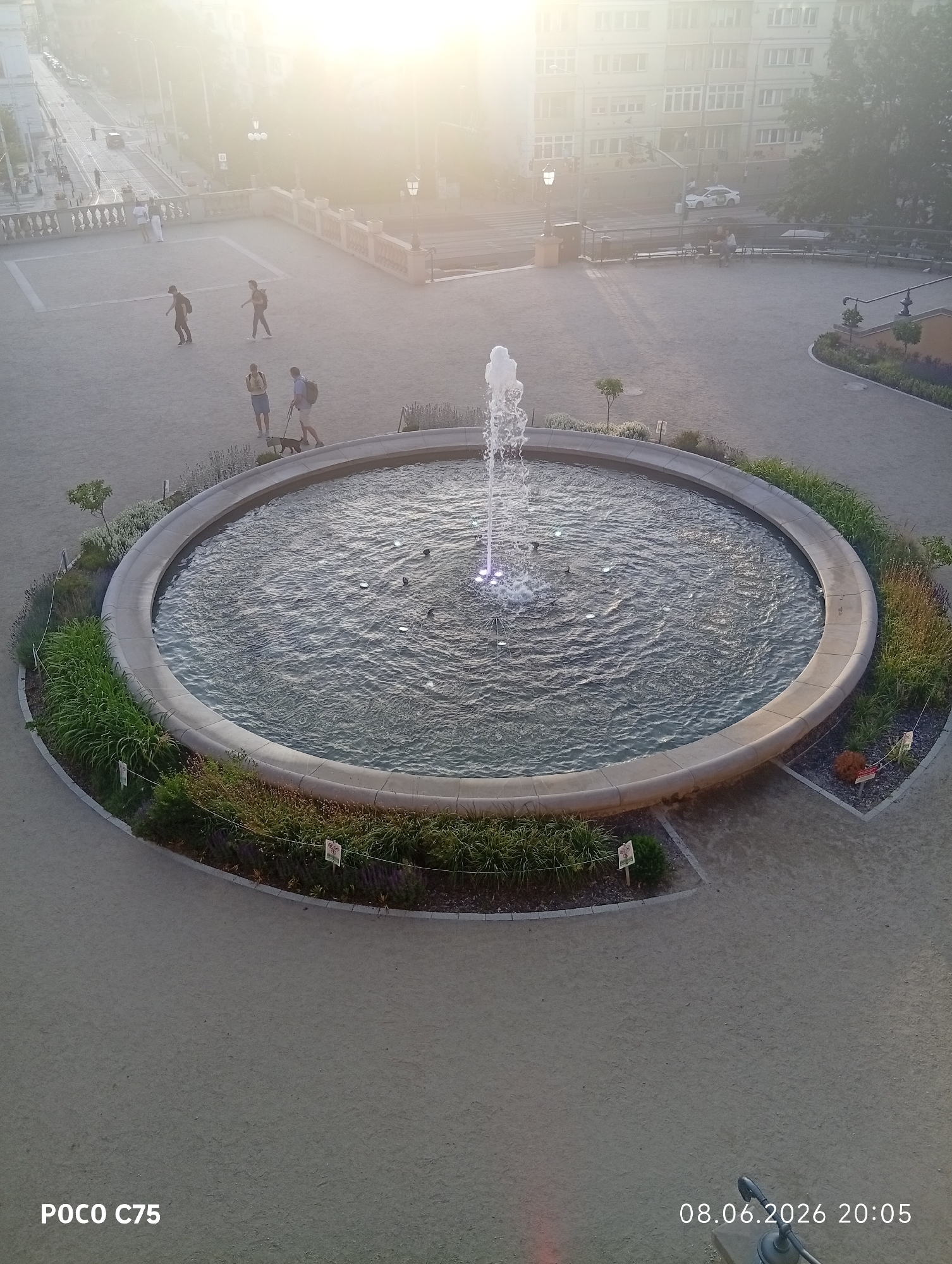 The image depicts a circular fountain in a public square, likely in Wrocław, Poland, given the timestamp format which is unusual for Polish dates. The fountain is active with water jets. In the background, a street with a tram line is visible, along with buildings and trees. Several people are present in the square: a group of three individuals are walking together near the street, and a man is walking a dog. The sun is shining brightly, causing a lens flare in the upper portion of the image. The ground of the square appears to be paved with gravel or small stones. The overall scene suggests a daytime setting, possibly late afternoon given the angle of the light.