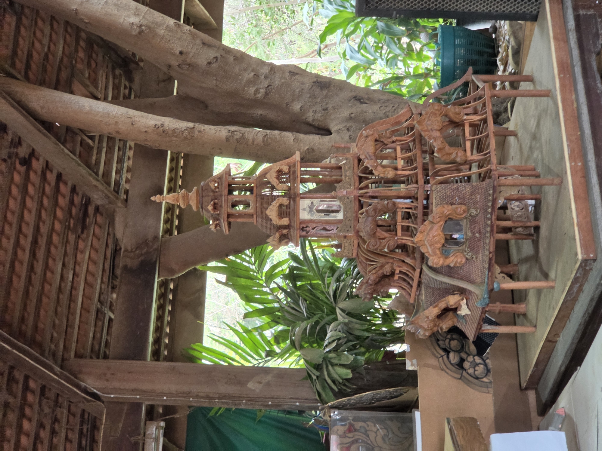 A detailed carving of a multi-tiered pagoda-like structure sits on a wooden frame within a building that incorporates tree branches into its architecture. The carving is intricate, with ornate details and possibly small windows or panels. The location is likely in Pattaya City, Thailand. The building’s construction features a wooden roof and support beams, with a large tree branch visible as part of the structure. In the background, greenery is visible through an opening, suggesting an outdoor or garden setting. The setting has the look of an open building or pavilion, blending nature and architecture.