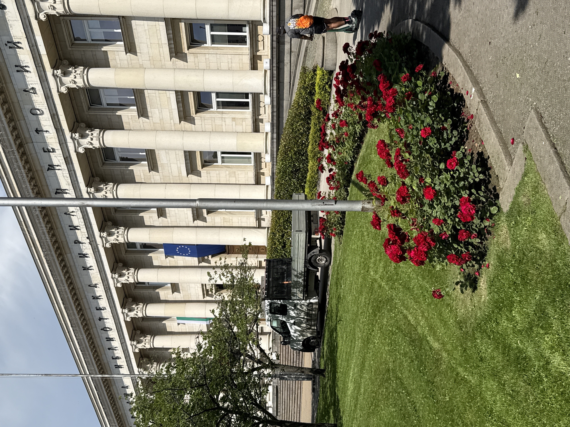 The image shows the National Library "Ivan Vazov" in Sofia, Bulgaria. The building's facade, adorned with classical columns and inscribed text, is visible. In front of the building, a well-maintained lawn with a vibrant bed of red roses is present. A white flatbed truck is parked on the left side of the image, partially obscured by trees. On the right side, a person is riding an electric scooter, wearing a dark t-shirt, shorts, a cap, and an orange backpack. The sky is partly cloudy, and the scene appears to be set on a sunny day.