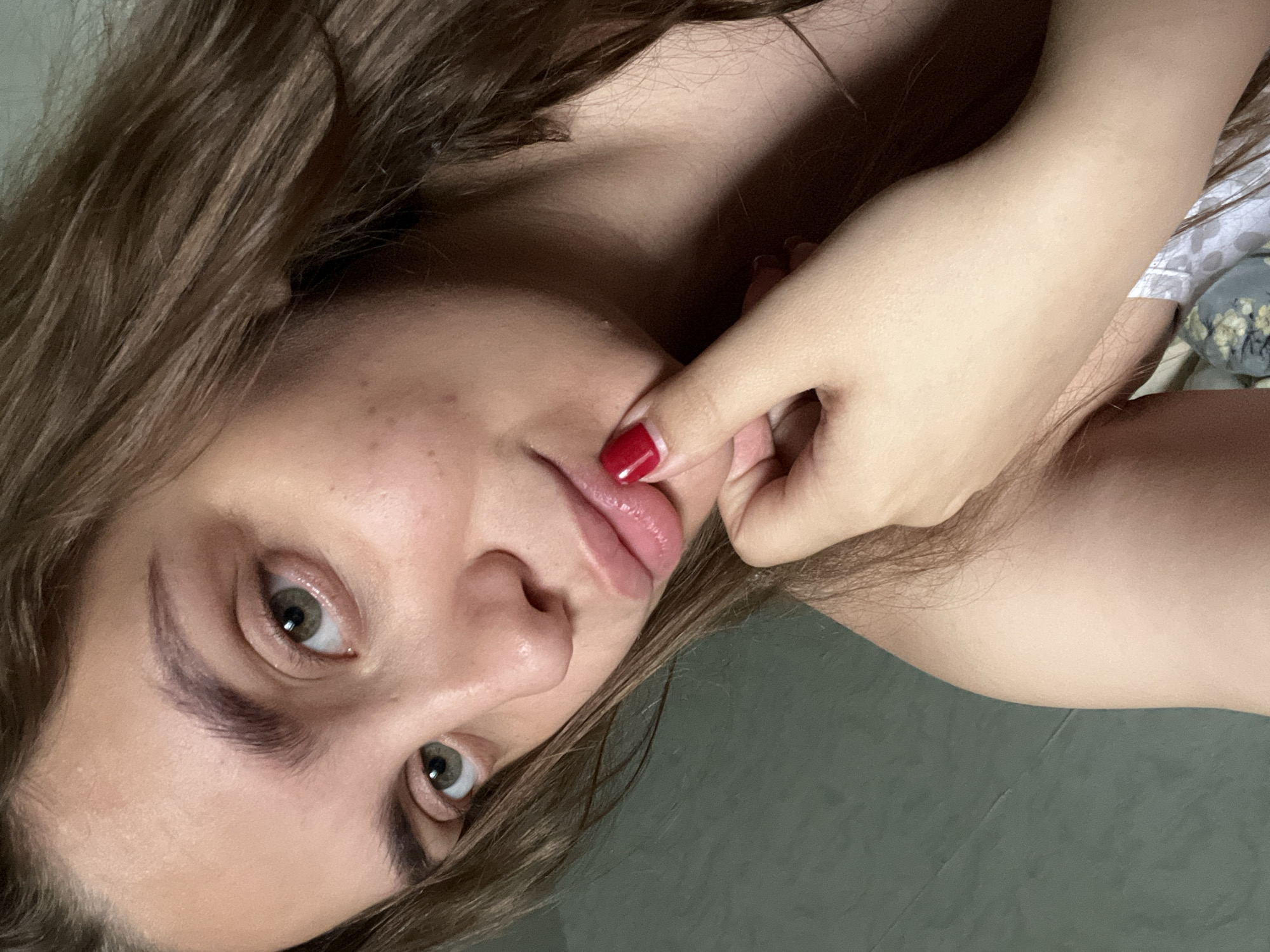 A close-up shot shows a young woman with light skin and brown hair. Her hazel eyes are open, looking directly at the viewer. She has full lips and a slightly upturned nose. Her left hand is near her face, with a finger gently touching her lower lip; the fingernail is painted red. The background appears to be a solid, dark grey-green surface, possibly a wall or floor. Other details of her clothing are not visible, however some details can be seen that look to be clothes near the top right edge of the image. The location of Chaikovsky, Russia is not discernable from the image.