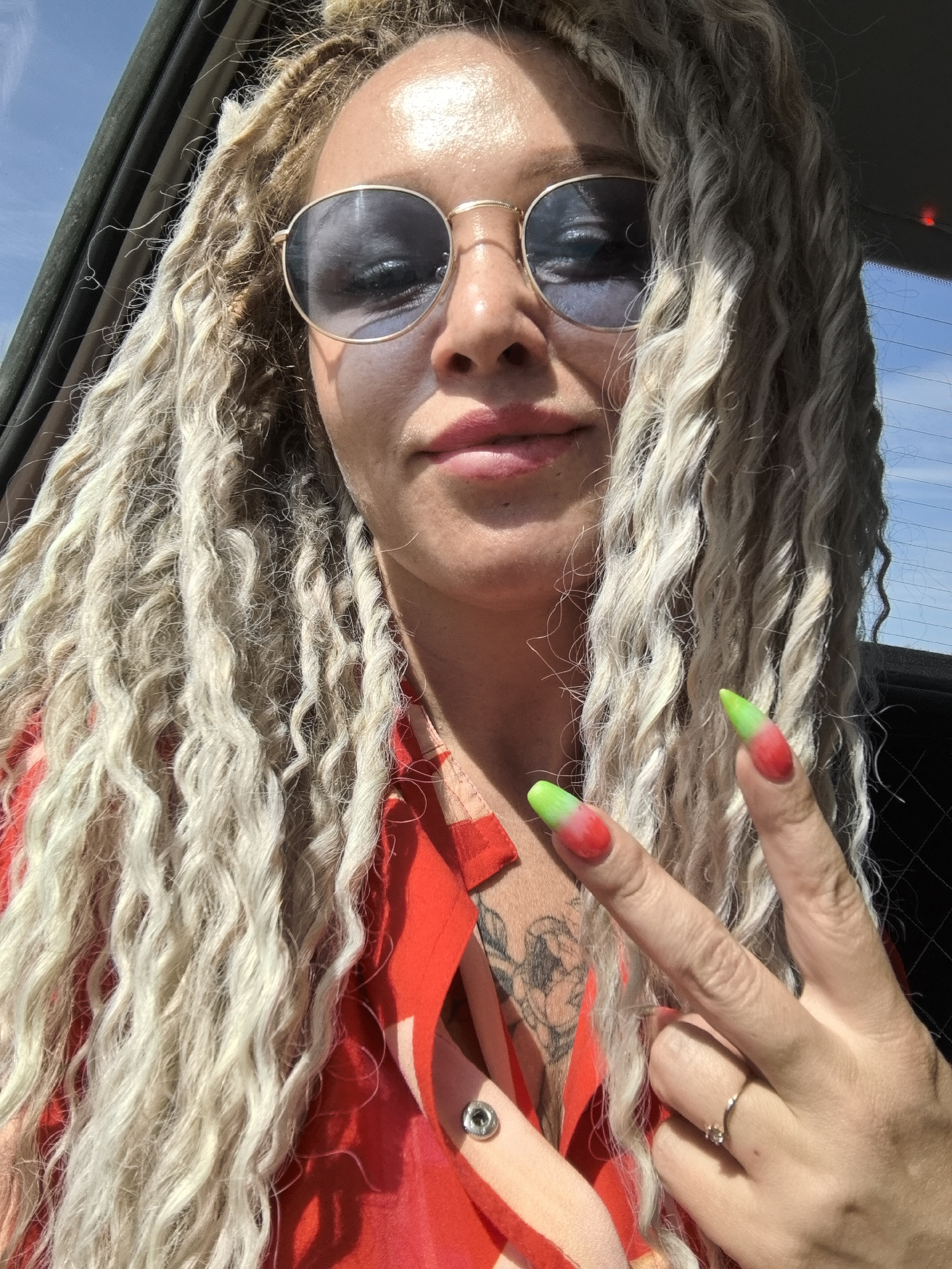 A medium shot of a woman with blonde dreadlocks and round sunglasses making a peace sign with her right hand. She is wearing a red patterned shirt with a button undone, revealing a tattoo on her chest. She is inside a car, as seen through the window. The location is not discernible from this image.