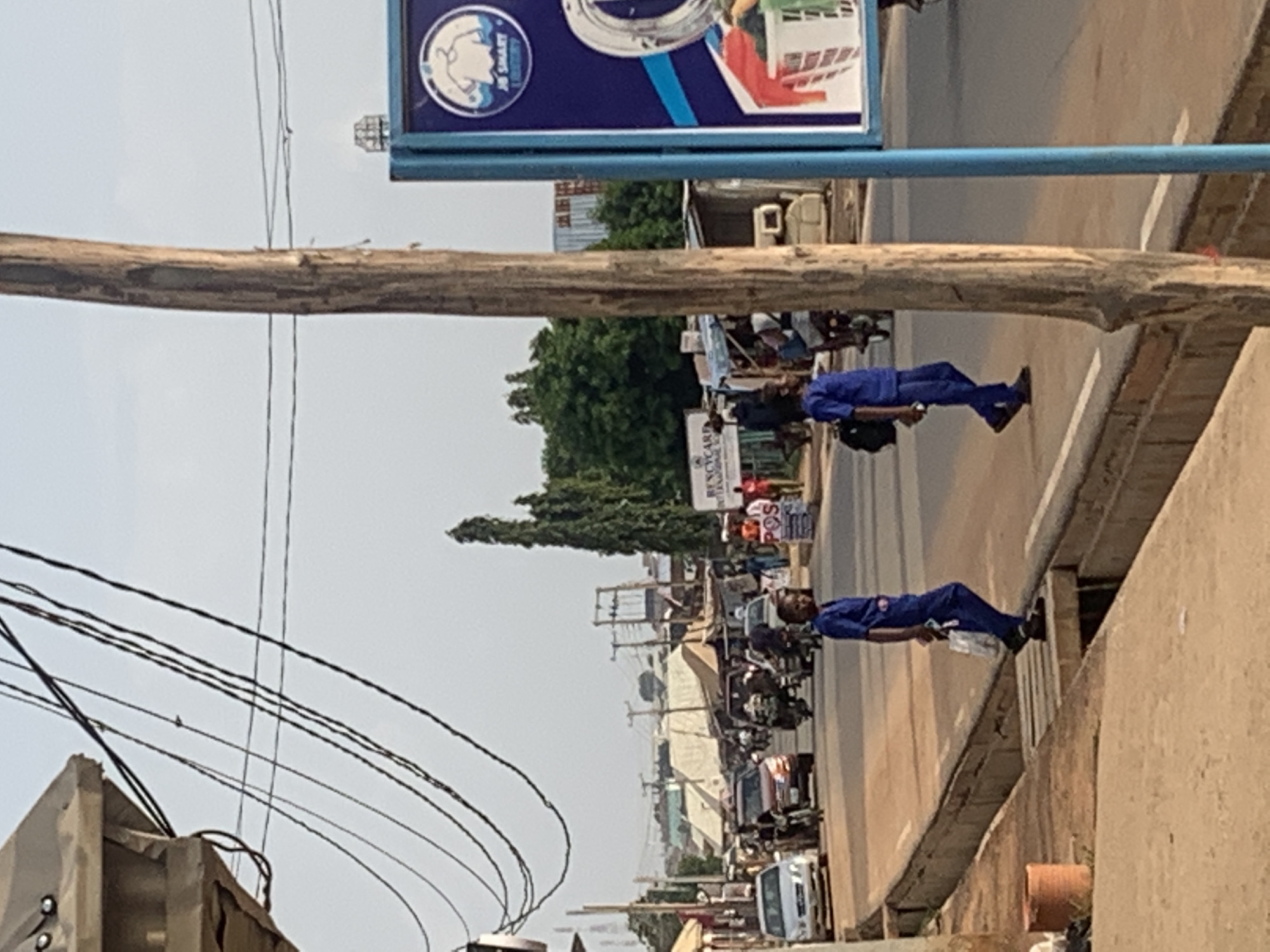 The image shows a street scene in Sabojibi, Nigeria. The street is lined with buildings and crowded with people, motorcycles, and vehicles. Two individuals in blue uniforms are visible on the right side of the frame. Power lines are overhead, and signage is present.