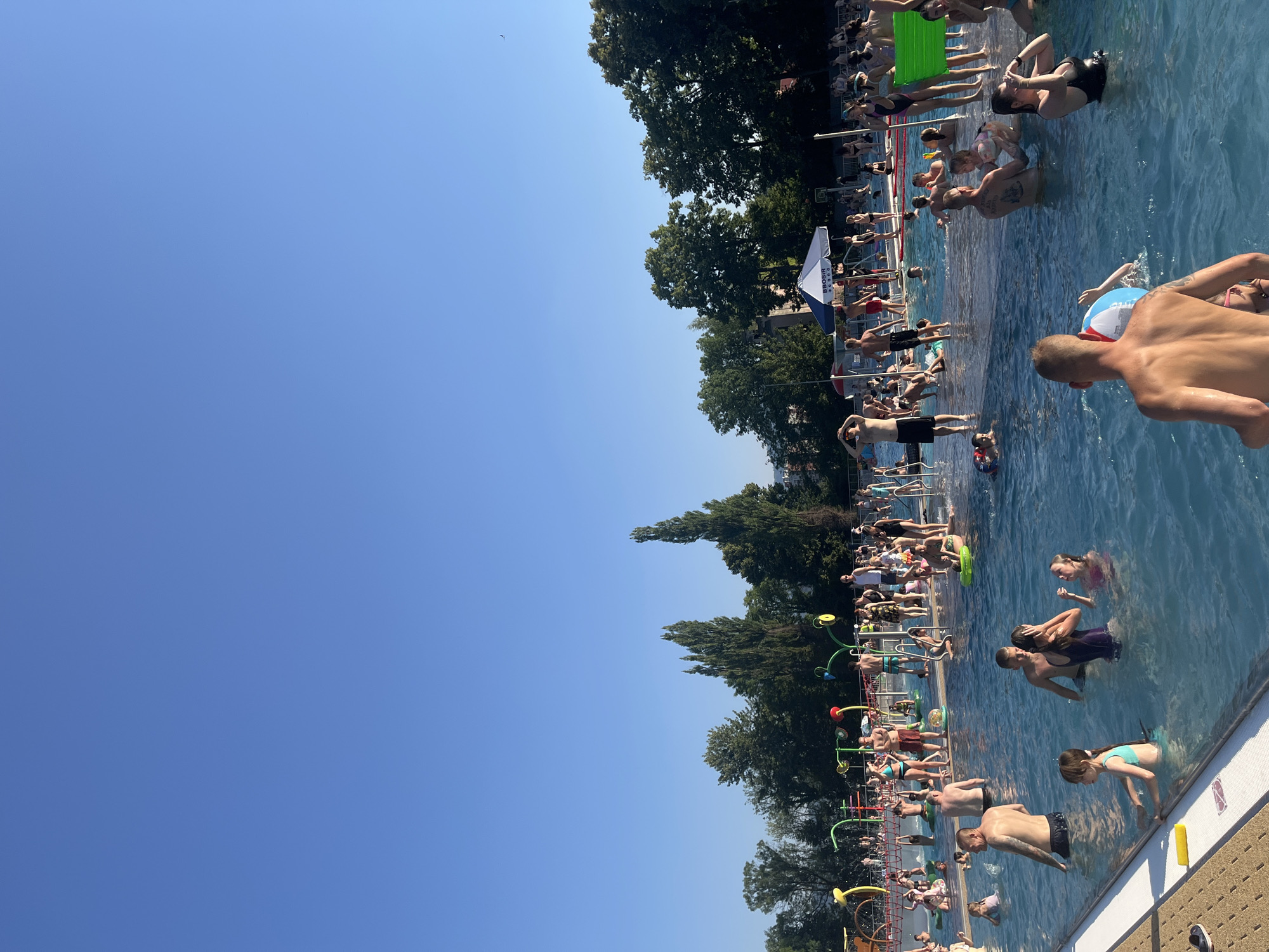 This media file depicts a crowded outdoor swimming pool on a sunny day. The pool is filled with people of all ages, engaged in various water activities such as swimming, splashing, and lounging. Some individuals are visible on the pool deck, either resting or preparing to enter the water. Children are also present, with some playing in the shallow end or interacting with water features. The scene is vibrant and lively, suggesting a recreational day out. The clear blue sky indicates good weather, likely during daylight hours, possibly mid-day or early afternoon, given the bright sunlight. The presence of trees surrounding the pool area suggests an outdoor recreational facility. A few objects are visible, including inflatable toys, pool ladders, and what appear to be water spray features in the background. One person is holding a red, white, and blue inflatable ball. The overall mood conveyed is one of enjoyment and relaxation. There is a partially visible banner with the text "BBQSBIR STO" on it, which may be related to the venue or an event.