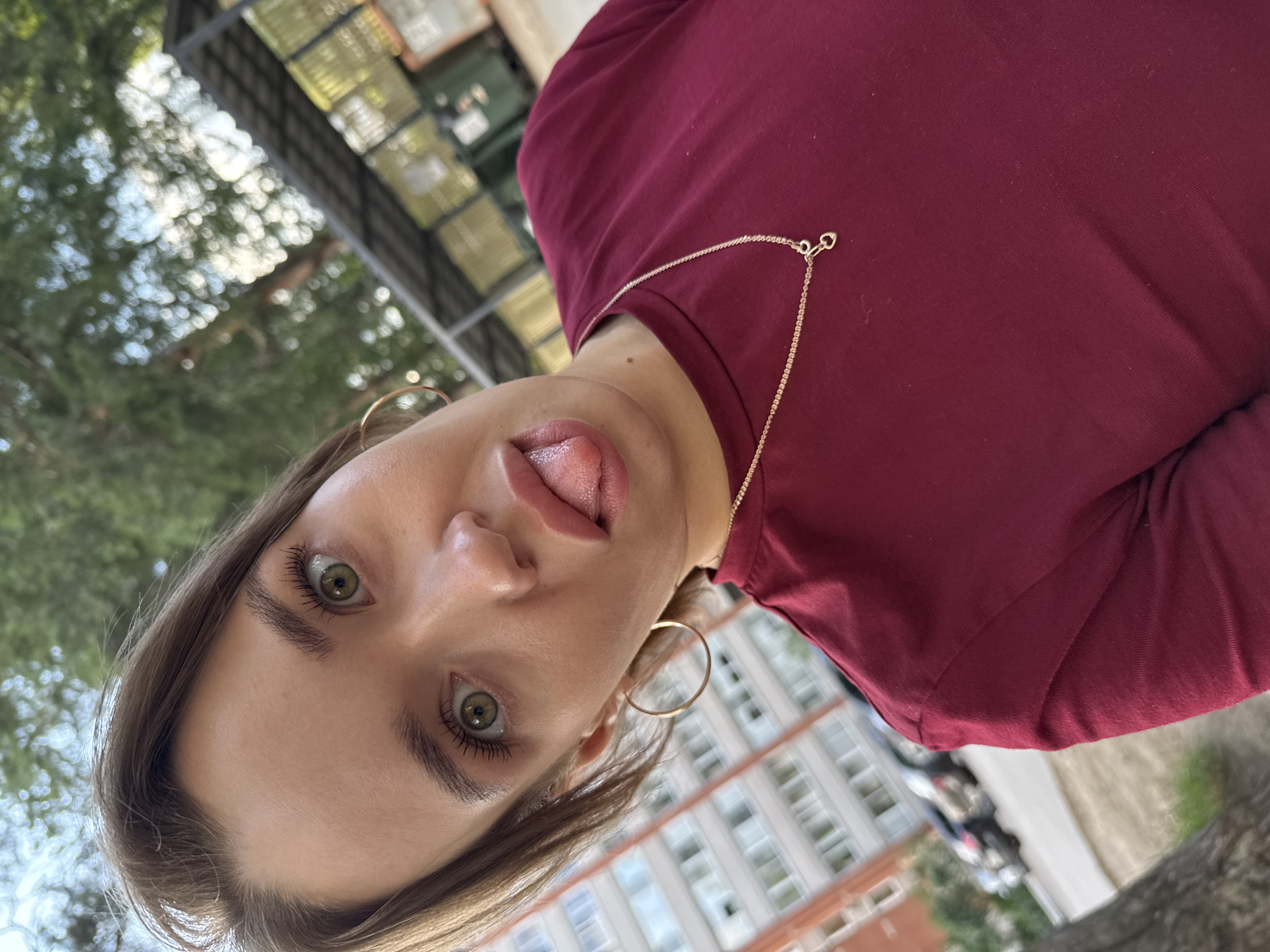 A young woman with green eyes and long brown hair is pictured in a close-up, low-angle shot, taken outdoors. She is wearing a burgundy top and a delicate gold chain necklace with a heart-shaped pendant. Her left eye is prominent, and her tongue is sticking out. Large hoop earrings adorn her ears. In the background, a blurred view of a modern building with numerous windows and trees suggests an urban or suburban setting. The lighting indicates it is daytime, possibly a bright, overcast day, given the soft light and lack of harsh shadows. The overall impression is playful and candid. The provided location context is Chaikovsky, Russia. There is no visible text in the image.