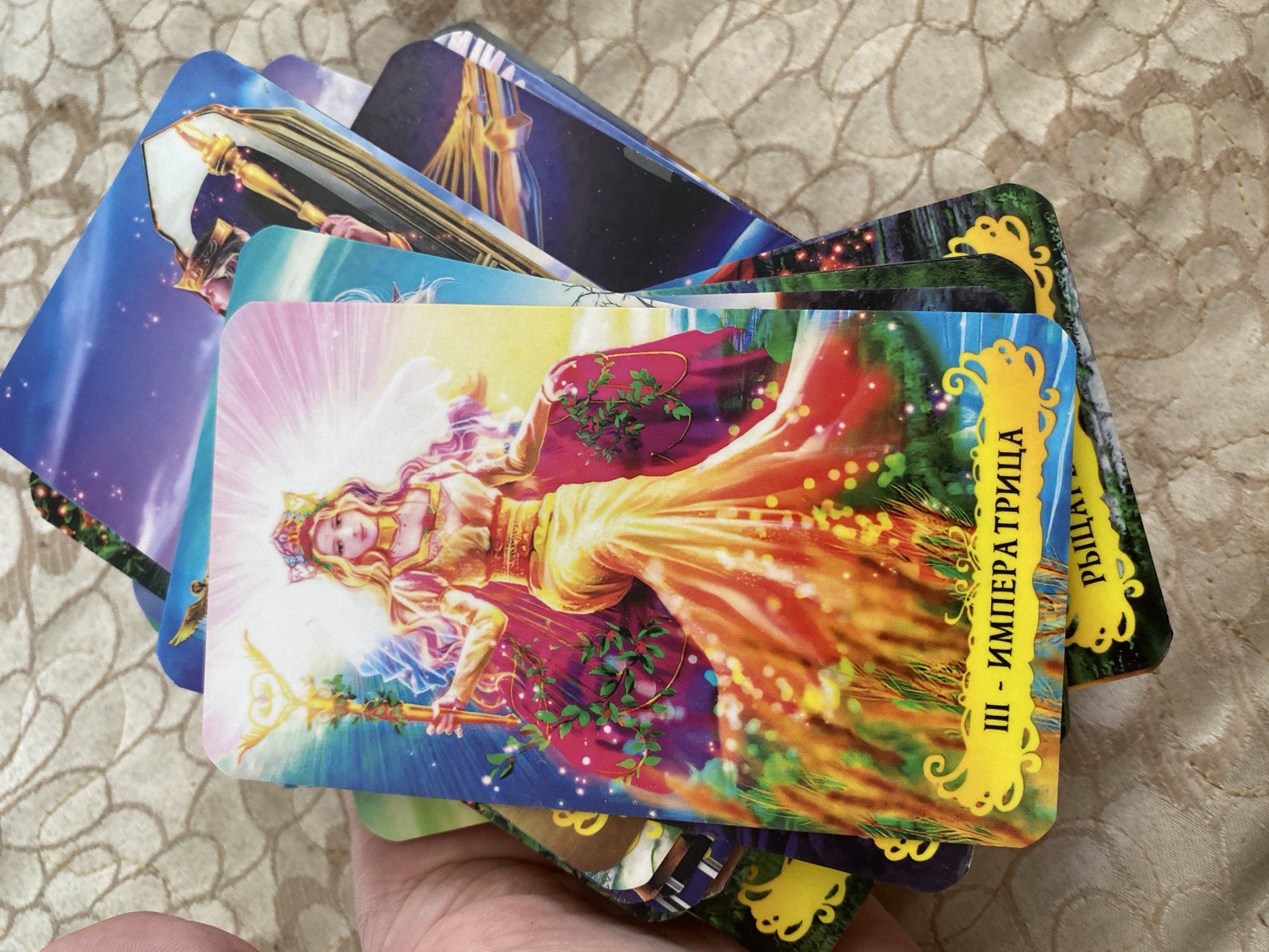 The image shows a close-up of a hand holding a stack of tarot cards. The top card features an illustration of a woman with a crown and a staff, dressed in a flowing golden gown. The card has Russian text at the bottom that reads "III - ИМПЕРАТРИЦА" (III - The Empress). Other cards are visible underneath, with various illustrated designs. The background is a patterned beige fabric. The photo was taken in Chornomorsk, Ukraine.