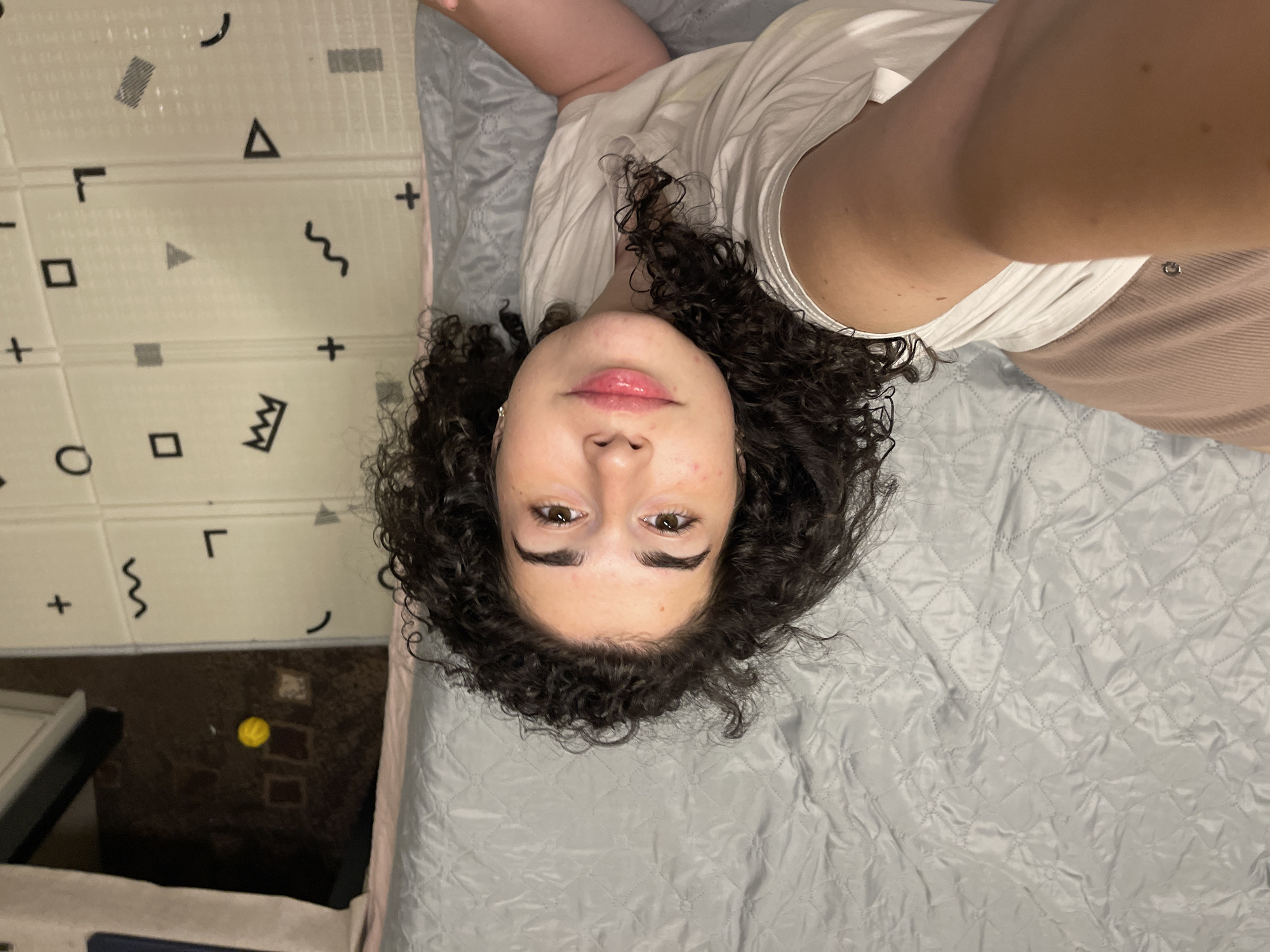 A young woman with dark, curly hair is lying on a bed in a room. She is wearing a white t-shirt and pink pajama pants. The bed is covered with a grey quilted comforter. In the background, there is a play mat with various geometric shapes. The lighting suggests it is nighttime, and the overall scene appears to be indoors, likely a bedroom. There is no visible indication of the location being in Sofia, Bulgaria.