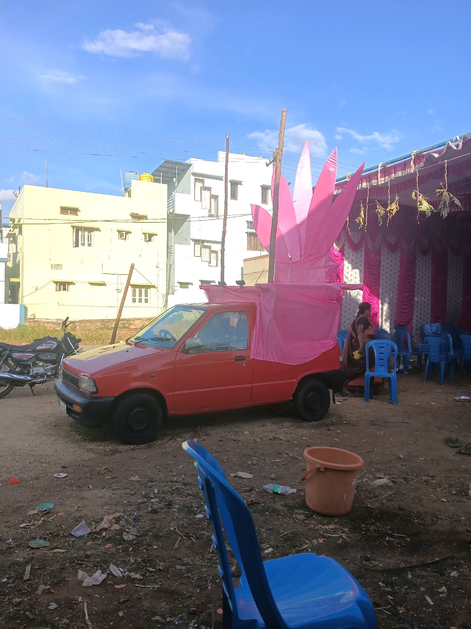 A red car, possibly a Maruti Suzuki Omni or a similar model, is parked on a dirt lot in Jagaluru, India. The car is decorated with a large, pink, petal-like structure, resembling a lotus flower, which is attached to its roof and extends upwards. The car is also draped in pink fabric. In the background, there are buildings, including a yellow one and a white one, and a blue sky with some clouds. To the right of the car, there is a temporary tent structure with pink and white drapes, and several blue plastic chairs are set up. A woman in a sari is standing near the car and chairs. A motorcycle is parked to the left of the car. The ground is covered with dirt, debris, and some scattered items like a plastic bottle and a bucket.