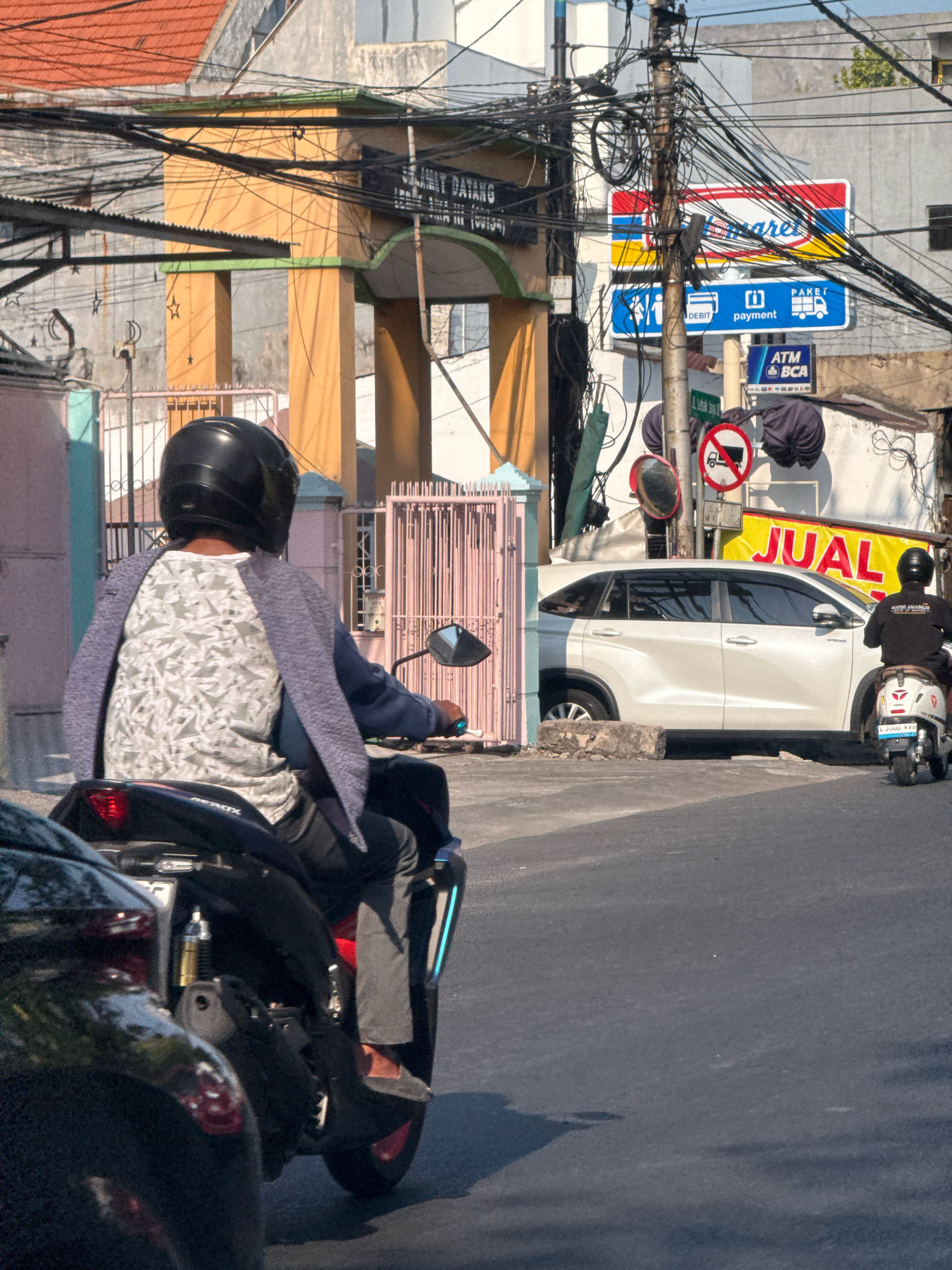 This is a street scene in Surabaya, Indonesia, under bright sunlight, with multiple power lines crisscrossing the sky and tangled around a utility pole. In the foreground, a person on a black motorcycle, wearing a black helmet and a light gray patterned shirt under a blueish-gray jacket, is riding away from the camera. To their right, a white SUV is parked on the side of the road. Behind the SUV, a sign reads "JUAL" in large yellow letters. Further back, there are buildings with orange and yellow accents, and a pink gated fence. A smaller white scooter is parked to the right of the SUV, with a person in a black shirt and helmet on it. Signs for "Indomaret" and ATM/BCA services are visible on the utility pole, along with traffic signs indicating "no trucks" and "no parking".