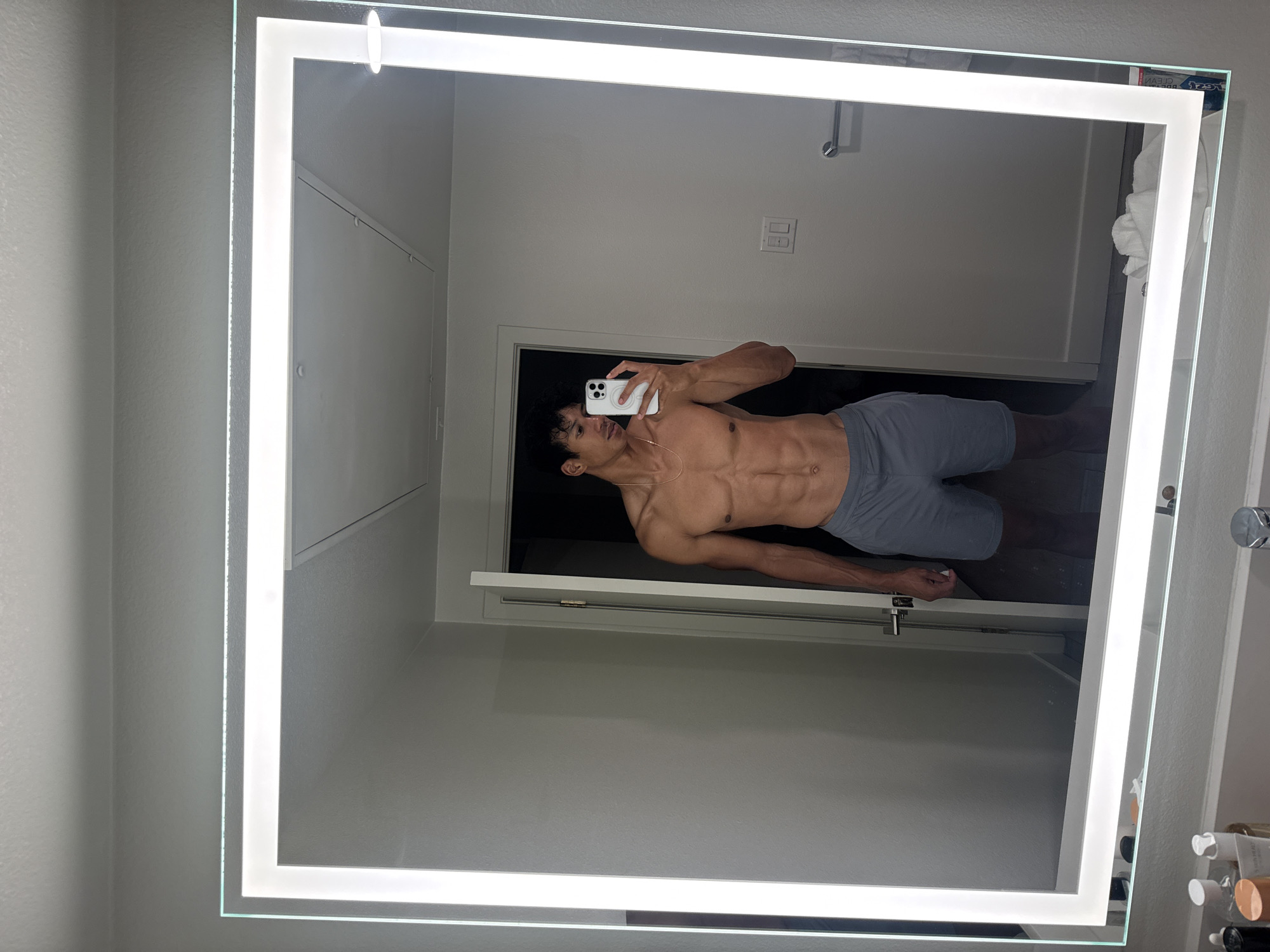 A shirtless man is reflected in an illuminated bathroom mirror. He is holding a white smartphone and appears to be taking a selfie. He has dark hair and is wearing grey athletic shorts. The bathroom is modern, with a light grey wall color, a white door, and light fixtures. The location is likely a residential bathroom in Los Angeles, United States of America.