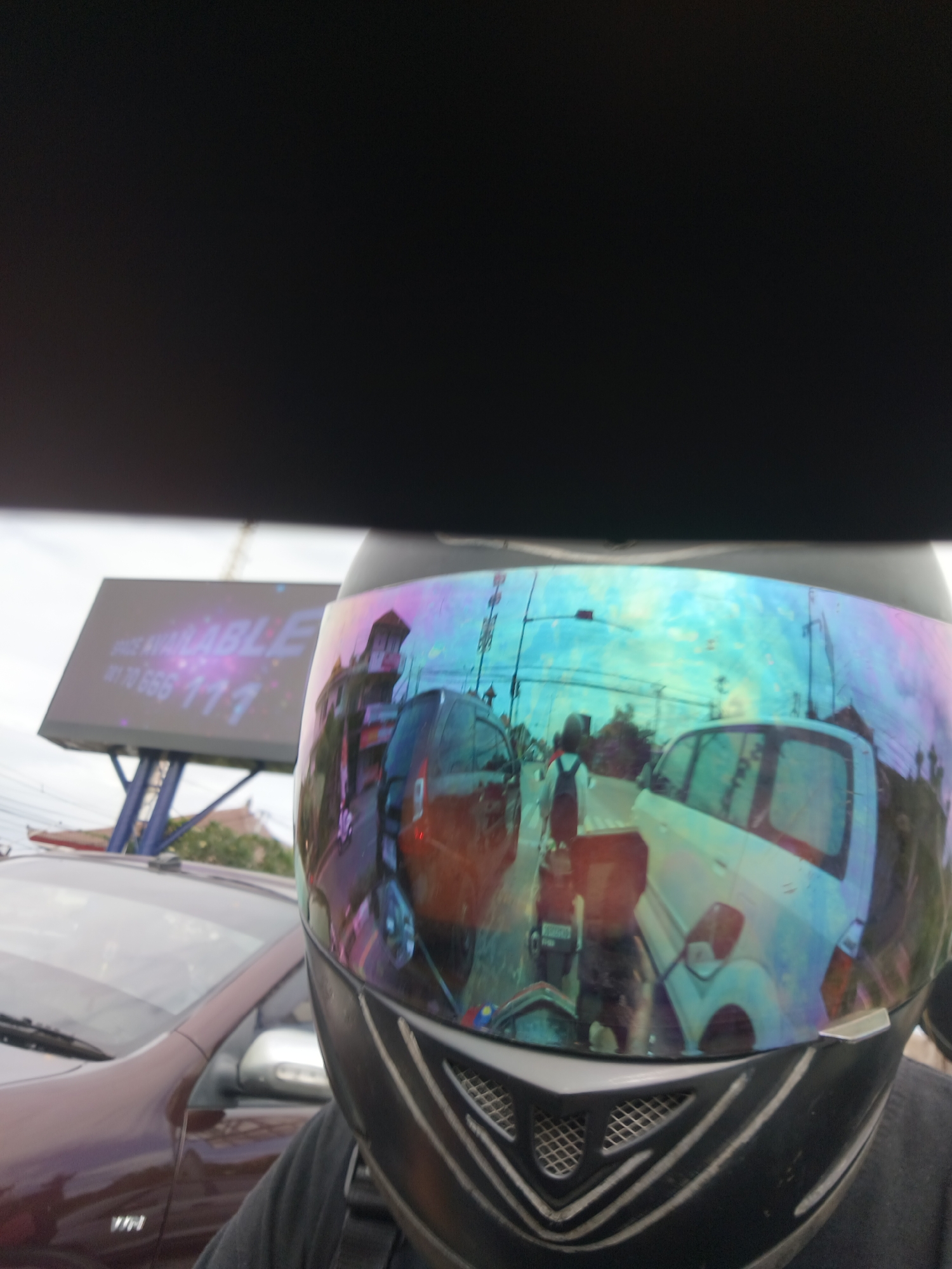 The image captures a close-up, point-of-view shot of a person wearing a helmet with a reflective visor. The visor reflects a busy street scene, likely in Denpasar, Indonesia, given the presence of a billboard that reads "SPACE AVAILABLE" and a phone number. The reflection shows traffic, including cars and motorcycles, as well as buildings and a traffic light. The billboard is positioned on a tall structure with blue supports, and trees are visible in the background. The foreground is dominated by the helmet, with part of a dark shirt visible on the wearer. The lighting suggests it is daytime, possibly overcast or during a time of day with diffused light, as indicated by the reflections on the visor and the general illumination of the scene.