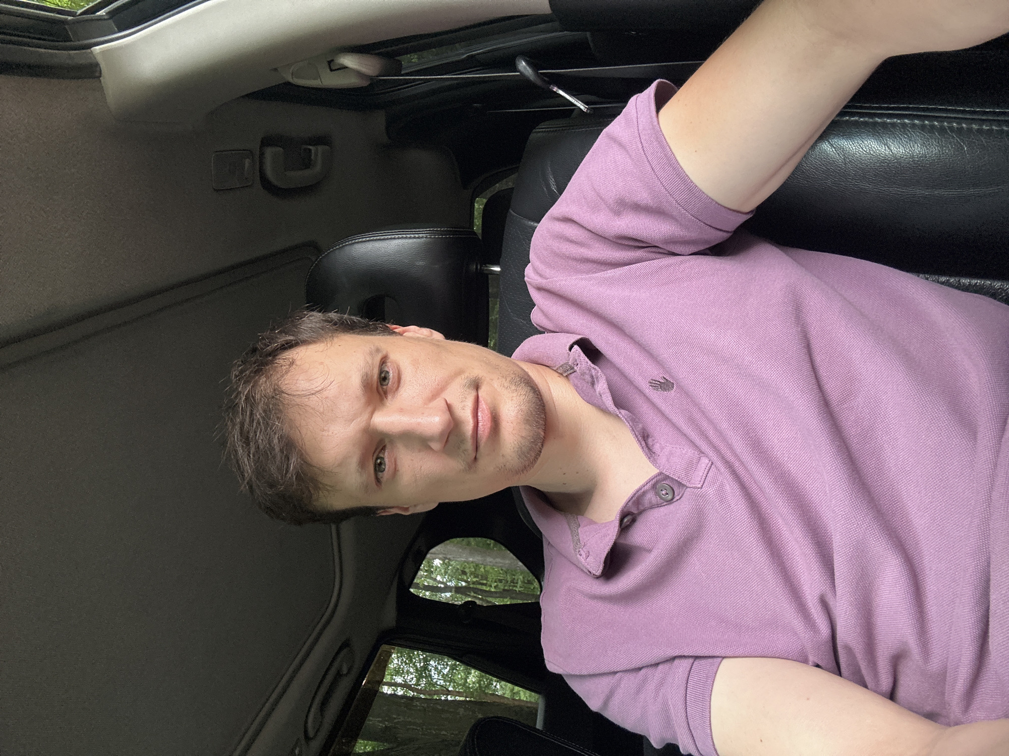 A man is sitting in the driver's seat of a car. He is looking directly at the camera. He has short, brown hair and is wearing a purple polo shirt. The car's interior is visible, including the headrest and part of the dashboard. Outside the car, trees and greenery can be seen through the windows. The image appears to have been taken during the daytime. There is no indication of the location being Shatava, Ukraine.