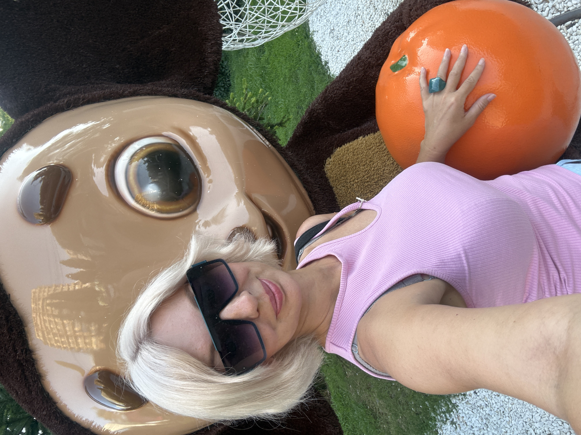 In Sochi, Russia, a woman with blonde hair and sunglasses poses for a selfie in front of a large, cartoonish brown character and an oversized orange. The woman is wearing a pink tank top and a blue ring on her right hand, which rests on the orange. Behind them, a reflection of a building can be seen on the character's shiny face, and grass and white gravel are visible on the ground.