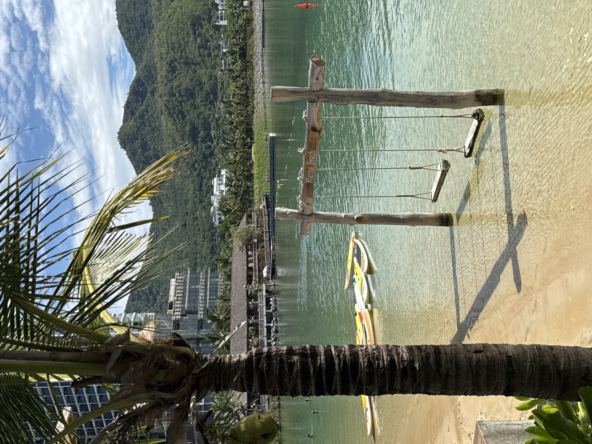 A serene coastal scene unfolds under a partly cloudy sky in Nha Trang, Vietnam. In the foreground, a textured palm tree trunk dominates the left side of the frame, with its fronds casting shadows on the sandy beach. A solitary green coconut hangs from one of the fronds. Adjacent to the beach, a calm, clear turquoise body of water reflects the sky and surrounding landscape.
Submerged in the shallow water are two wooden swings, each with two ropes suspending a simple wooden seat. The swings are anchored by thick, weathered wooden posts planted in the sand beneath the water's surface. A wooden sign with the inscription "Marina Beach" is affixed to the crossbeam connecting the swing posts.
Several paddleboards and kayaks are neatly arranged on the sandy shore, some partially submerged in the water, suggesting recreational activities. In the middle ground, a thatched-roof building with wooden walkways extends over the water, appearing to be a restaurant or resort facility. Further in the distance, several people can be seen swimming and wading in the water.
The backdrop features lush green mountains that rise majestically, dotted with buildings and palm trees, indicating a well-developed coastal area. Modern high-rise buildings are also visible on the left side of the frame, contrasting with the natural beauty of the mountains and the tropical foliage. The overall atmosphere is tranquil and inviting, capturing the essence of a beachfront resort.