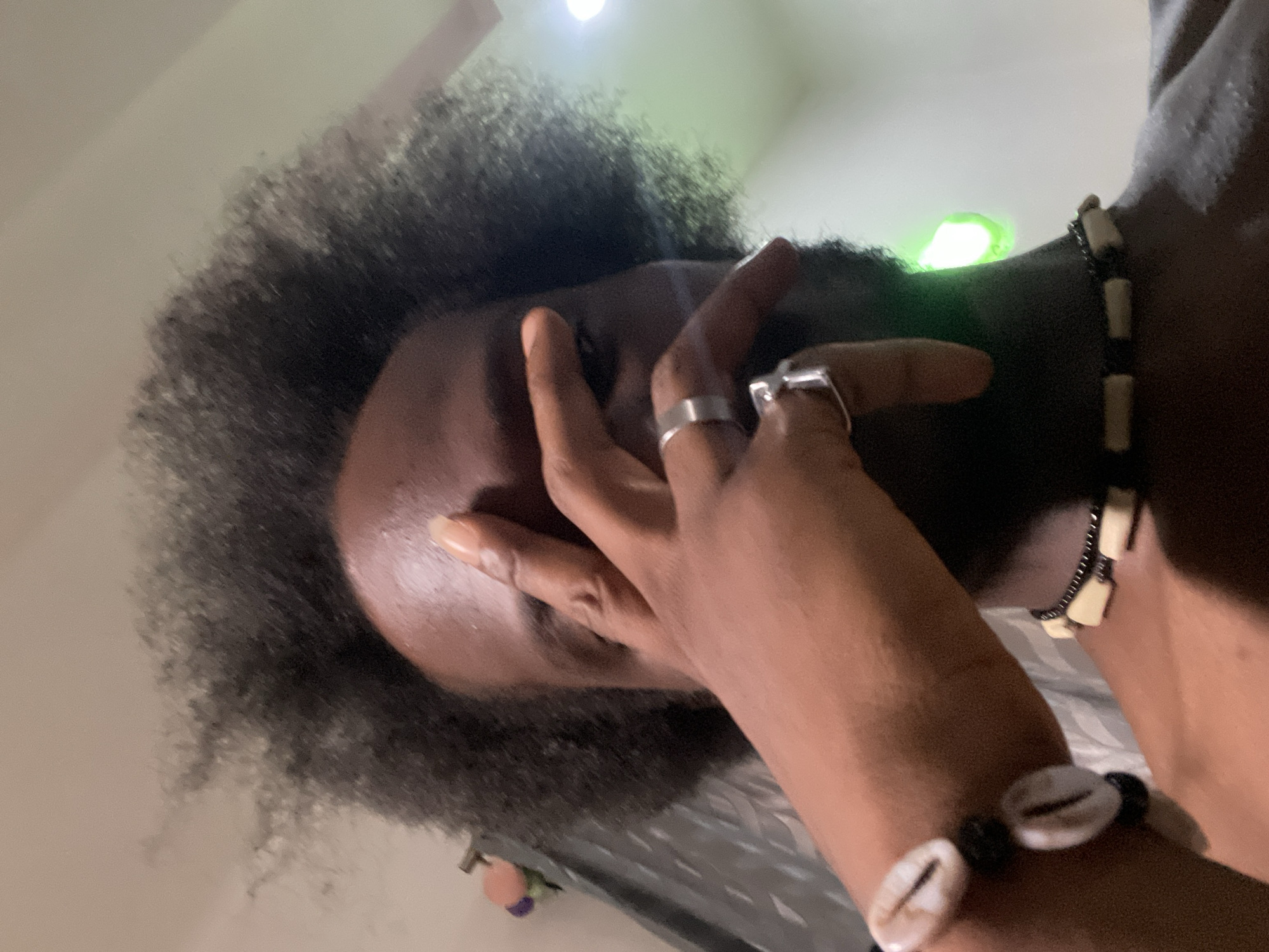 A close-up, low-angle shot depicts a dark-skinned individual with afro-textured hair covering their left eye. The subject is wearing multiple rings on their fingers and a bracelet with cowrie shells. A beaded necklace is visible around their neck. The environment is lit by a light source above, which is reflected on the forehead of the individual. The location, Kuchingoro, Nigeria, is not discernible from this image alone.