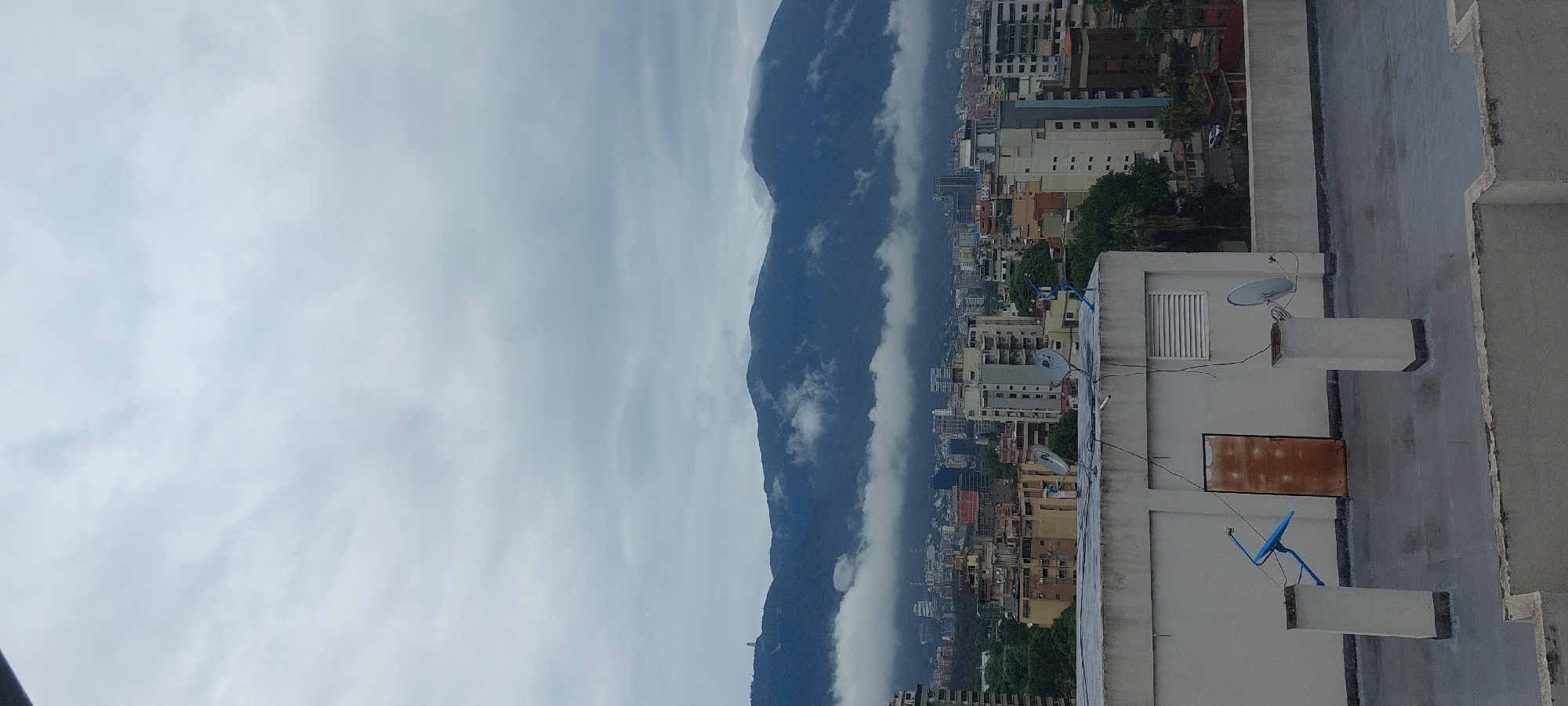 The image shows a cityscape of Caracas, Venezuela, taken from a high vantage point, possibly a rooftop. A mountain range is visible in the background, partially obscured by clouds. In the foreground, the rooftops of buildings are visible, including a white building with a satellite dish. The sky is overcast.