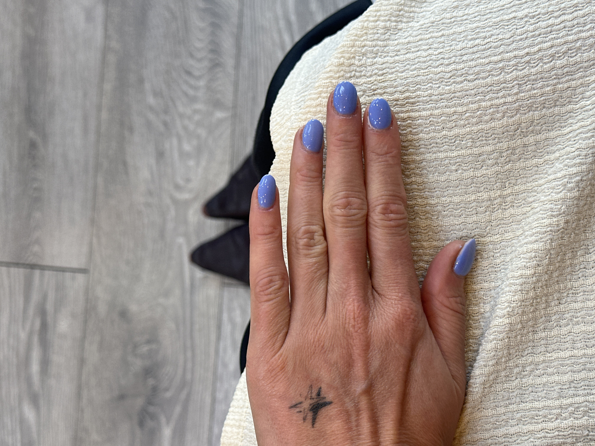 A hand rests on a cream-colored fabric with a textured, wavy pattern. The hand has short, oval-shaped fingernails painted a light blue-purple color with subtle shimmer. There is a small, faded tattoo of a star-like symbol on the back of the hand, near the wrist. To the left of the hand, there is a glimpse of a gray, wood-patterned floor and part of a black object (possibly a shoe or bag). The location is not discernible from the image content.