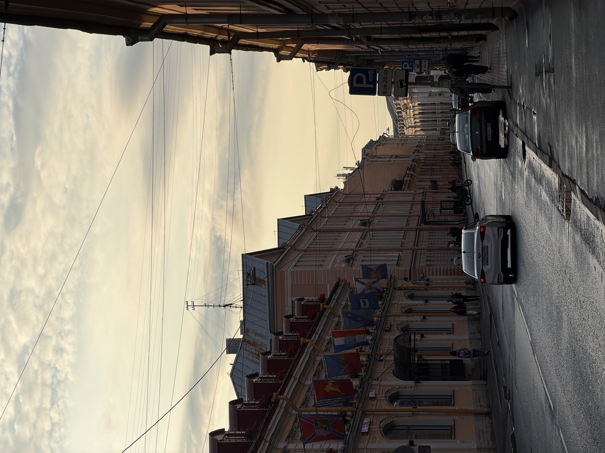 This is a vertical, medium-shot, eye-level photograph of a street in Saint Petersburg, Russia. The street is paved with asphalt and has a wet sheen, indicating recent rain or damp conditions. The sky is overcast with light, wispy clouds. Overhead, numerous electrical wires crisscross the sky, supported by poles and attached to buildings.
On the left side of the street, a long, three-story building features a light orange facade and a mansard roof with red dormer windows. Several flags, hung vertically from the building's facade, display various emblems and crests. People are visible walking on the sidewalk in front of this building.
On the right side of the street, another building with a darker facade runs parallel to the street. A sidewalk runs along the front of this building. A traffic sign with a blue "P" for parking is visible, along with other traffic regulatory signs. Several people are walking on this sidewalk, and a few cars are parked along the curb.
In the foreground, two cars are driving down the street. A white sedan is in the left lane, and a darker sedan is in the right lane. The streetlights cast long shadows, suggesting it is late afternoon or early evening. The overall atmosphere of the image is subdued and slightly melancholic, likely due to the overcast sky and wet pavement. The architecture and street signs are consistent with urban settings in Saint Petersburg.