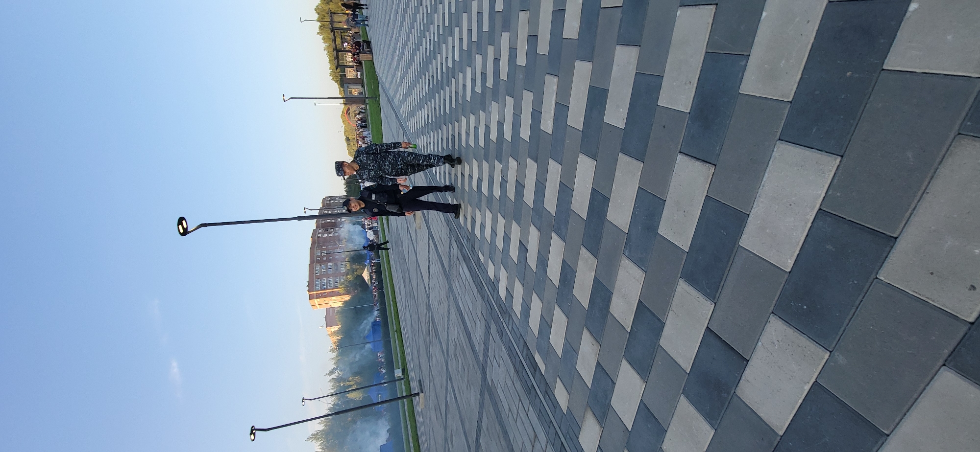 The image shows two uniformed men walking on a paved walkway. The man on the left wears a dark uniform with a badge on his chest and a black cap. The man on the right wears a camouflage uniform and a cap. They are walking towards the camera. The walkway is paved with rectangular blocks of alternating gray and dark blue colors. In the background, there are streetlights, trees, a brick building, and a crowd of people gathered in tents. Smoke is visible in the middle ground, possibly from a performance or an event. The sky is clear and blue, indicating daytime. The scene appears to be in an outdoor public space, possibly a park or a festival area, in the city of Oskemen, Kazakhstan.