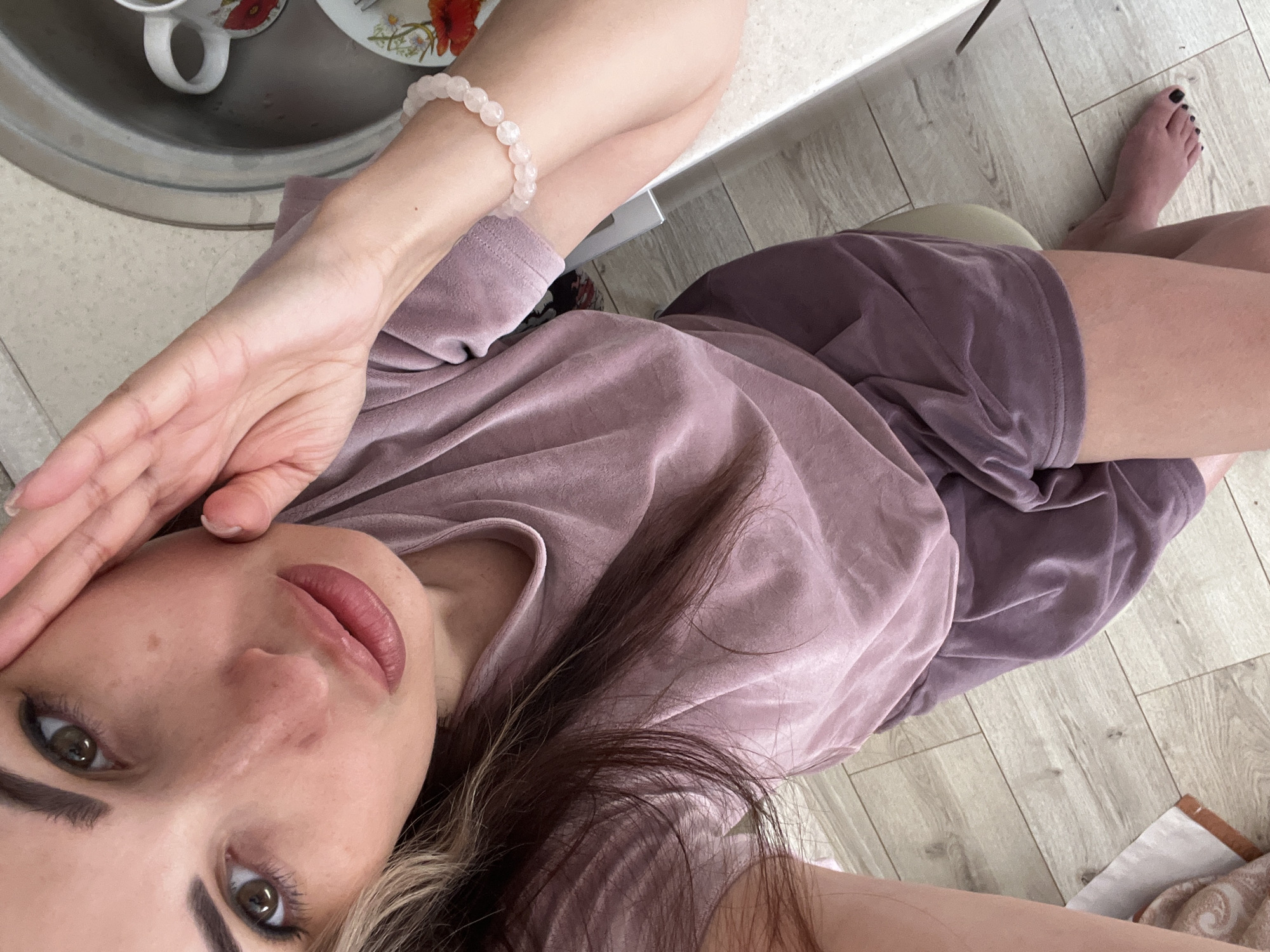 A medium shot captures a woman from the chest up, angled downwards. She has long, dark brown hair with blonde highlights, and is looking directly at the camera with her right hand resting on her cheek. She is wearing a dusty rose velour t-shirt and matching shorts. A light pink beaded bracelet is visible on her left wrist. The background shows a light-colored countertop with a stainless steel sink and a white mug with a floral design. The floor appears to be light-colored wood planks.