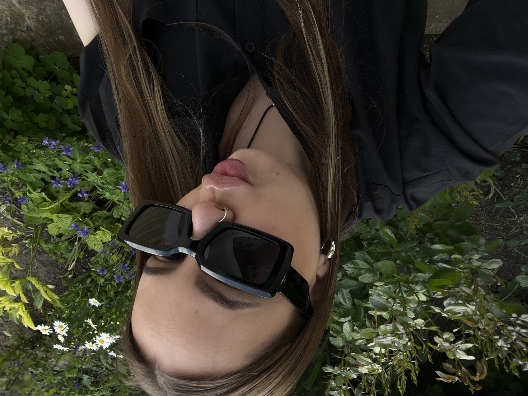 The image is a close-up, angled portrait of a young woman, taken outdoors in Novopokrovskaya, Russia. The photo is rotated 90 degrees clockwise, showing her head tilted back and angled upwards, filling a significant portion of the frame.
The woman has long, straight brown hair with lighter highlights, which cascades around her face and extends towards the upper left of the image. She is wearing a black collared shirt, unbuttoned at the neckline, revealing a thin black necklace or choker. Her eyes are obscured by stylish black rectangular sunglasses. A silver nose ring is visible on her right nostril, and a silver hoop earring adorns her left ear. Her lips are slightly parted, conveying a relaxed and casual expression.
The background consists of a lush outdoor garden setting. In the upper left portion of the image, amidst vibrant green foliage, small clusters of purple flowers and white daisies are visible. The upper right part of the background features denser green bushes and leaves. The scene is illuminated by soft, natural daylight, indicating that the photo was taken during the day, possibly under an overcast sky or in the shade, contributing to a tranquil and serene atmosphere. There is no visible text in the image.