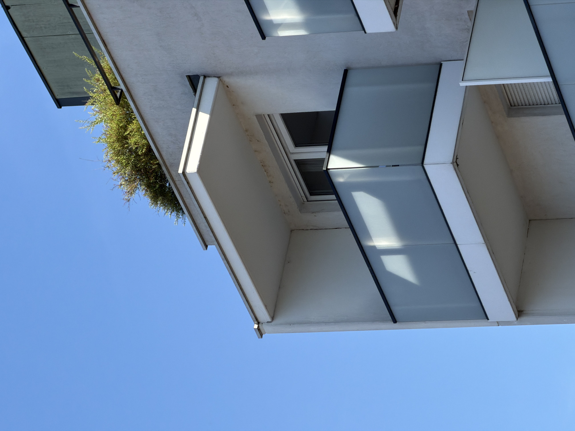 This is a close-up, low-angle, outdoor, eye-level shot of a modern building's exterior on a clear, sunny day. The dominant colors are the bright blue sky and the off-white stucco of the building.
The building features multiple balconies with frosted glass railings. The main balcony in the foreground is white with a white soffit and a dark gray trim. Behind it, a window with white trim is visible. Above this balcony, another, smaller balcony or overhang is present, also white. On the roof of the building, a dense cluster of green foliage is visible, suggesting some form of rooftop planting or greenery. To the right of the main balcony, a section of another balcony with similar frosted glass railing and white trim is visible.
The architectural style appears contemporary and minimalist. There are no people or animals visible in the image. The overall impression is one of clean lines and modern design against a serene blue sky. The image does not provide any specific landmarks or indicators to suggest it is located in Strasbourg, France.