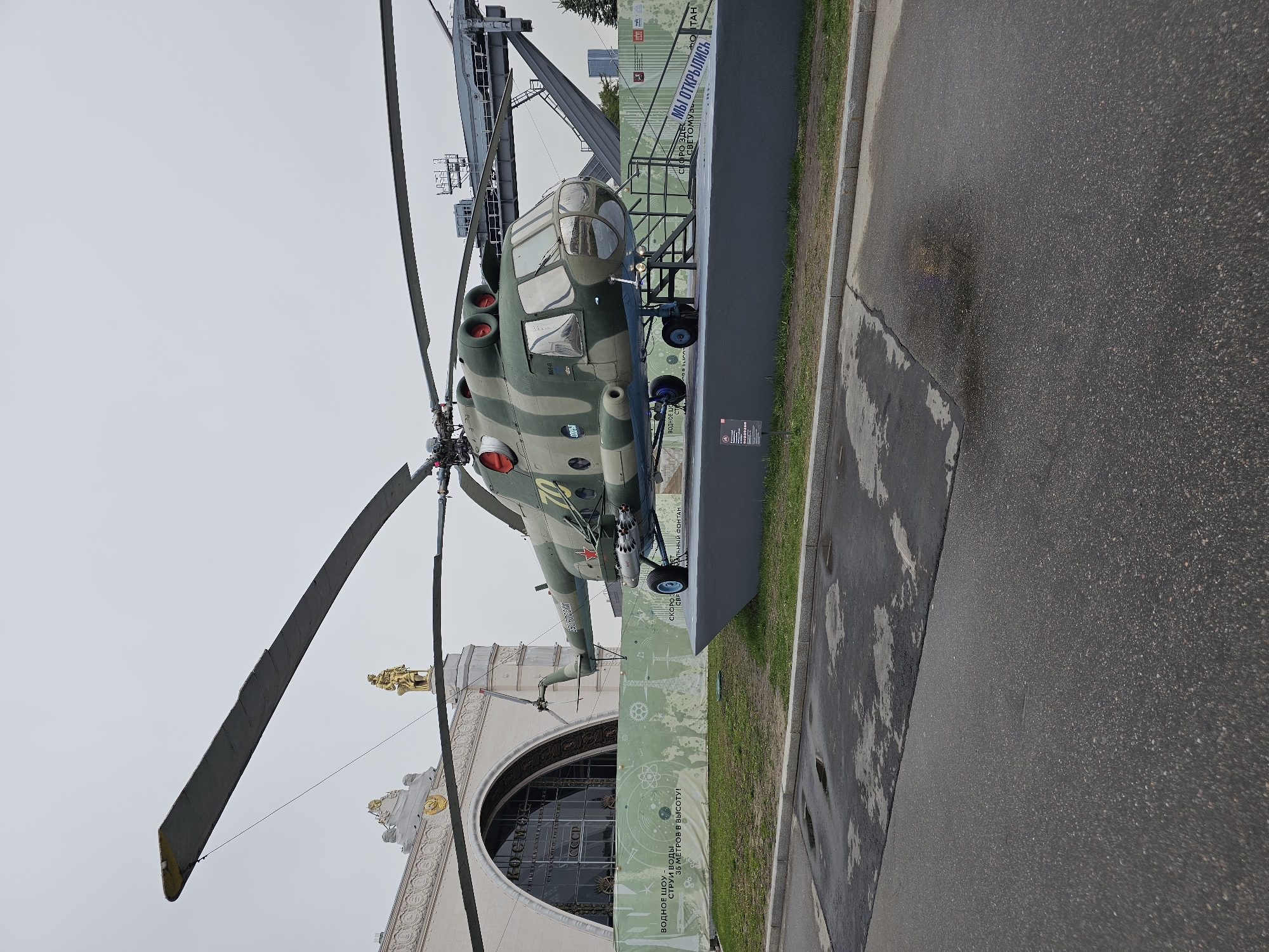 A military helicopter, painted in camouflage green and tan, is prominently displayed. The number "02" is visible on its side. It's mounted on a raised grey platform next to a wide sidewalk and a two-lane paved road. The setting is outdoors, with an overcast sky. A tall building is partially visible in the background, its architecture featuring decorative moldings and a golden statue on top. Russian writing is seen on a sign next to the helicopter. It is located at VDNKh in Moscow, Russia.