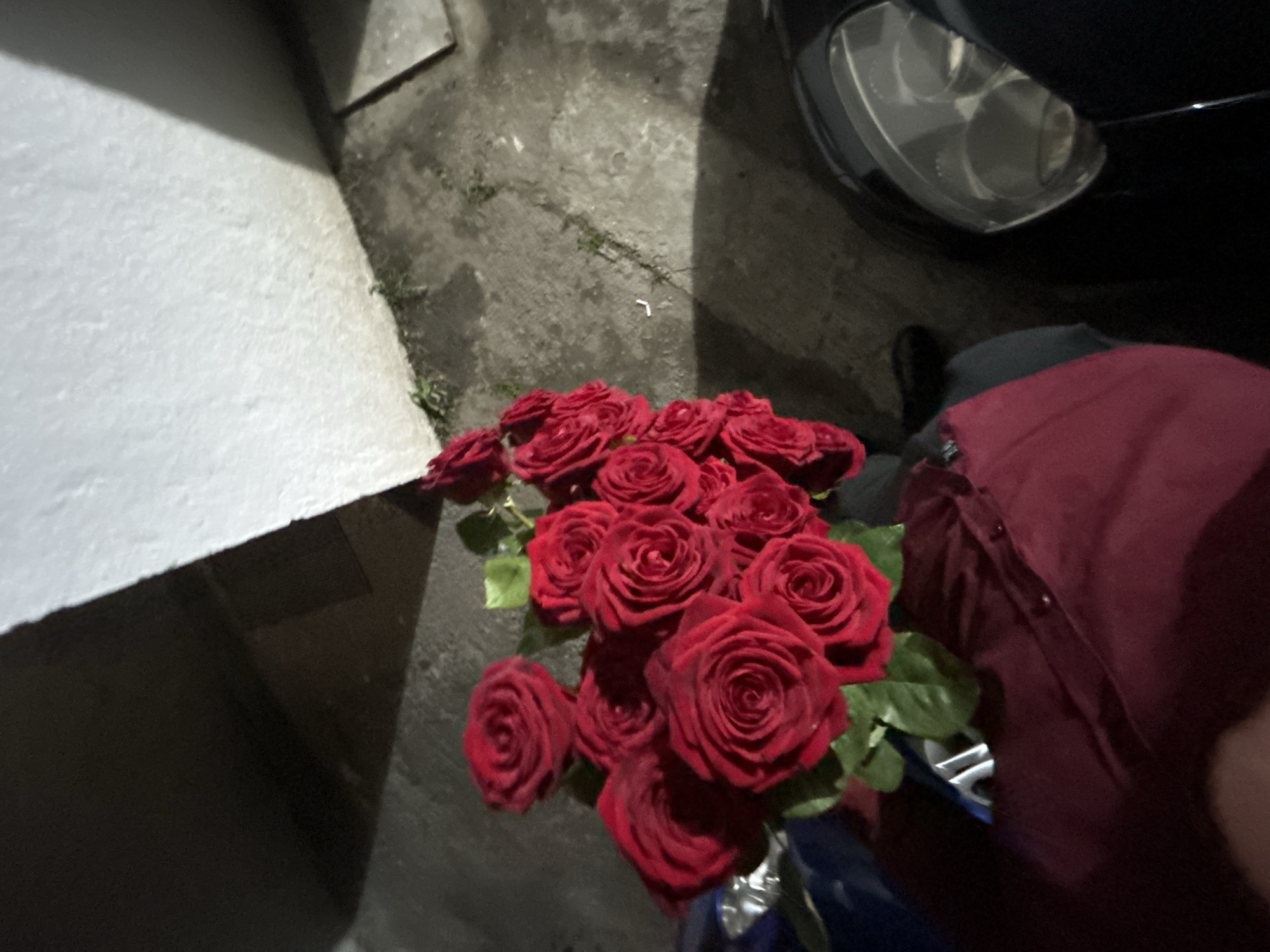 A bouquet of red roses rests on the ground near a person wearing a burgundy shirt and standing next to a car, likely taken at night due to the darkness and car's headlight in the top right corner. The location is possibly in a city based on the concrete ground, but there's insufficient data to pinpoint the location within Zlatograd, Bulgaria. The bouquet is the central object, with a focus on the arrangement of the roses. No specific actions or interactions are observable beyond the placement of the roses on the ground.