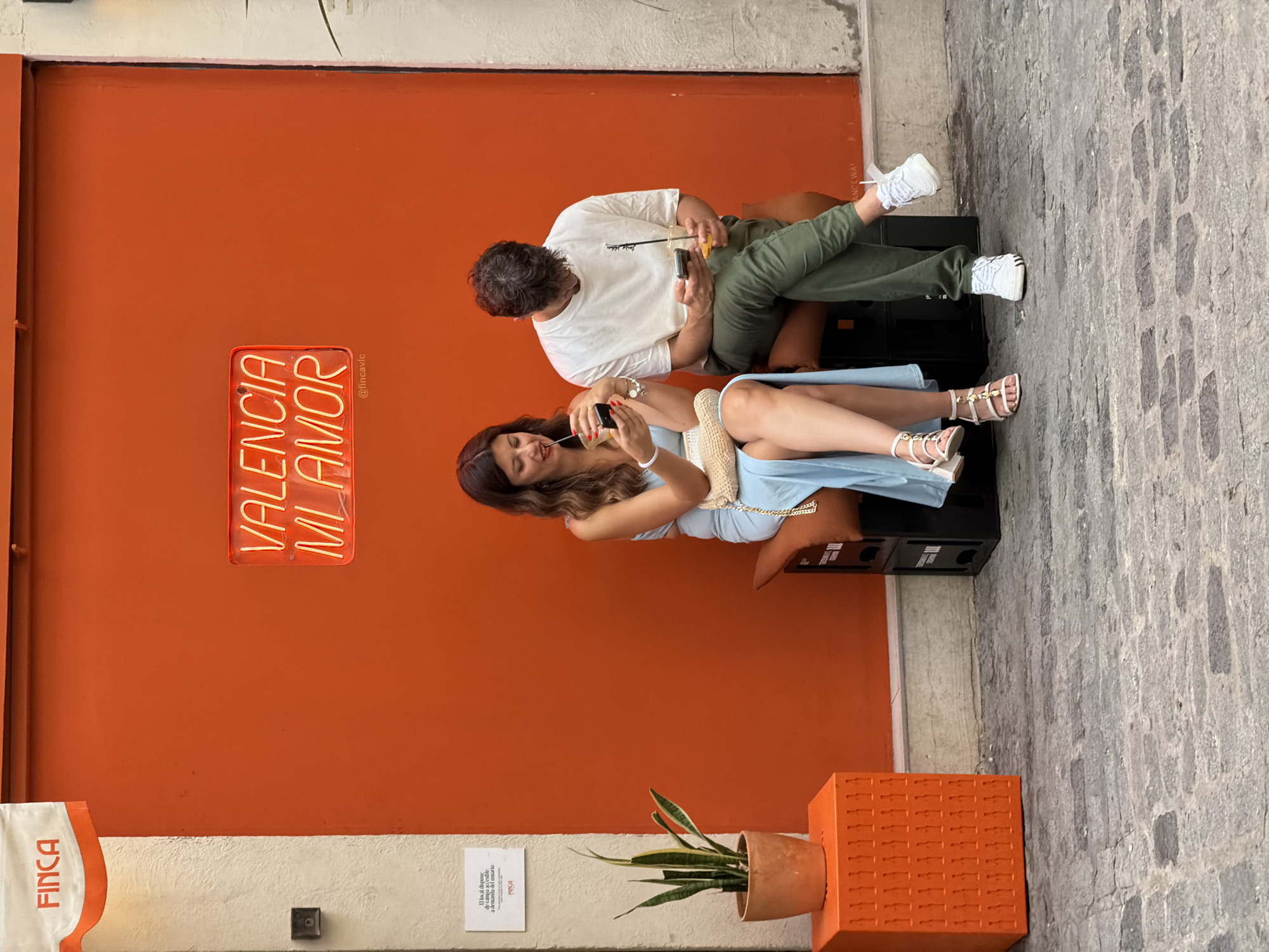 A man and a woman are seated side by side on black crates in front of an orange wall. The woman, on the left, wears a light blue dress and is looking at her phone while holding a drink. The man, on the right, wears a white t-shirt and green pants, also looking at his phone and holding a drink. A neon sign on the wall above them reads "VALENCIA MI AMOR". To the left of the couple, a potted plant sits on an orange decorative cube, and a white sign is affixed to a cream-colored wall. The ground is paved with cobblestones.