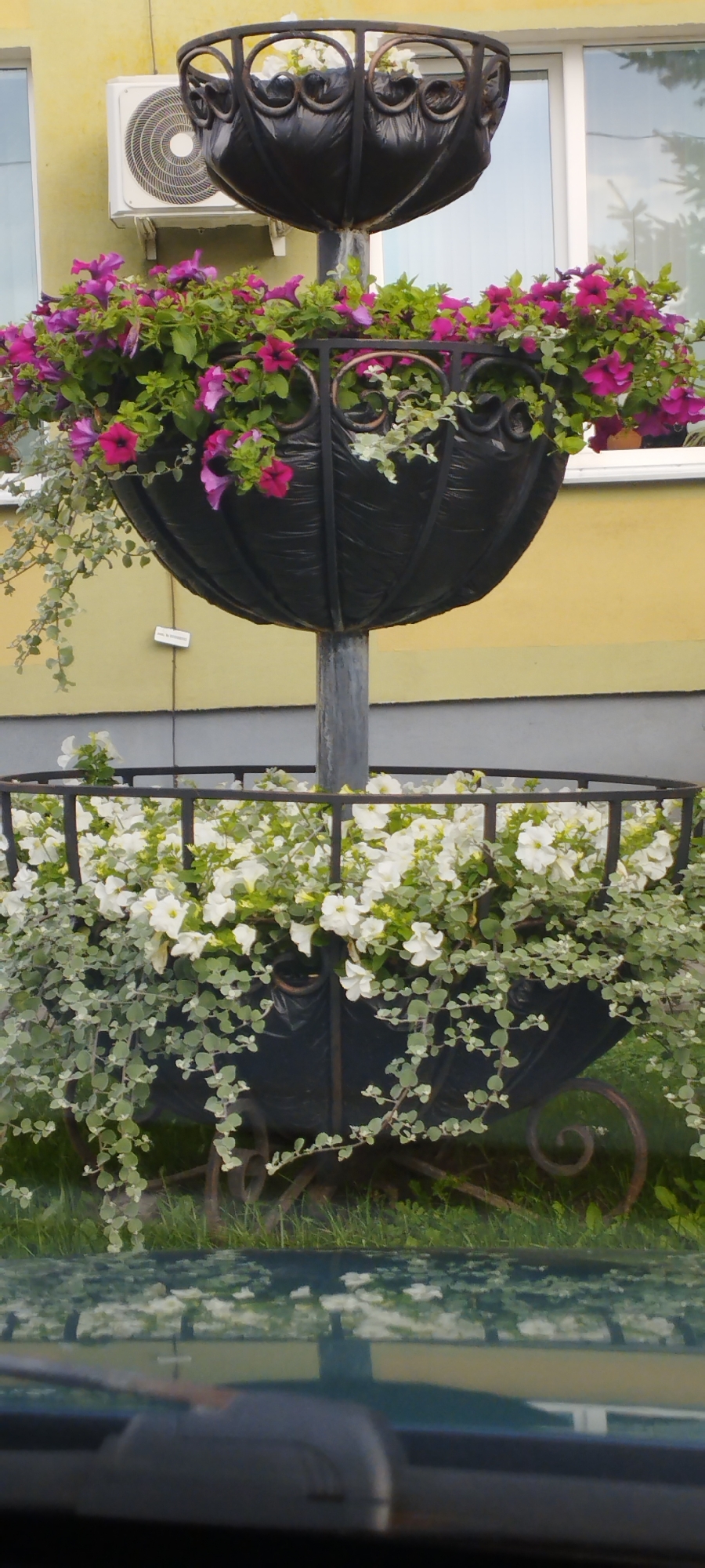 This is a vertical, eye-level, medium shot of a two-tiered planter filled with flowers. The top planter is a large, dark, urn-shaped container overflowing with vibrant pink and purple petunias and some trailing green foliage. The bottom planter is a wider, rectangular, dark container also filled with white flowers and similar trailing green foliage. Both planters are made of black metal with decorative scrollwork and are supported by a central dark metal pole. Behind the planters is a yellow building with a white window and an air conditioning unit mounted on the wall. The ground beneath the planters is covered in green grass and some of the planters' ornate metal bases are visible. A blurred reflection of the scene is visible in the foreground, suggesting the shot was taken from inside a vehicle. The location is Unknown, Unknown.