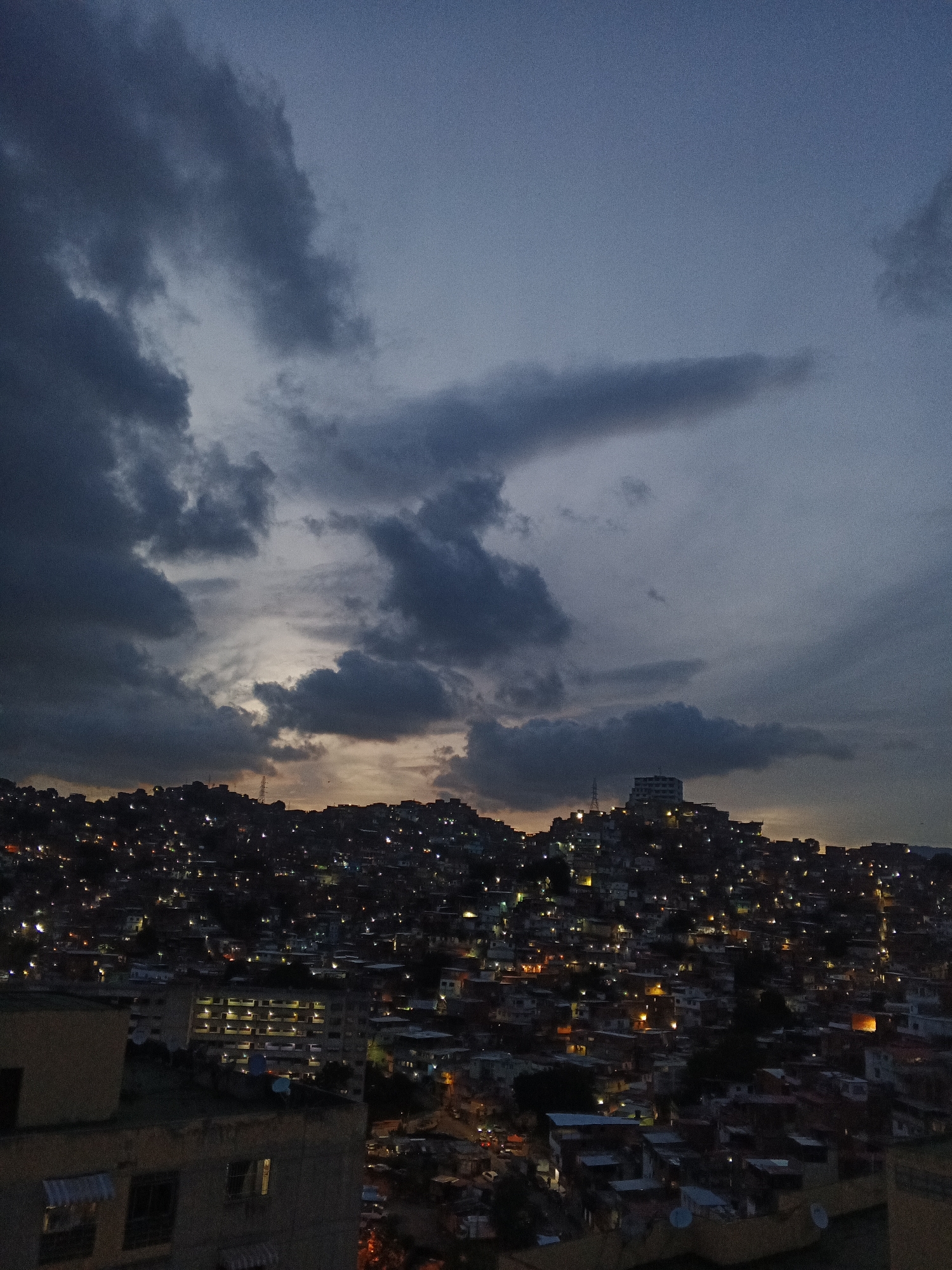 This image depicts a sprawling hillside urban landscape at dusk or early night in Caracas, Venezuela. The upper two-thirds of the frame are occupied by a deep blue-grey sky heavily laden with dark, uneven clouds. Hints of orange and yellow light from a setting sun or distant source peek through the clouds on the horizon, illuminating the undersides of some clouds. The lower third, extending up the steep hillsides, reveals a dense mosaic of residential buildings, characteristic of a *barrio* or informal settlement. Countless small, warm-toned lights emanate from windows and structures, creating a glittering pattern across the dark terrain. A larger, multi-story building is visible on the crest of a prominent hill towards the center-right. In the immediate foreground, parts of other buildings are visible, showing windows with interior lights and awnings. No individual people, specific actions, or interactions are discernible. The scene is static, focusing on the illuminated urban environment under a dramatic sky.