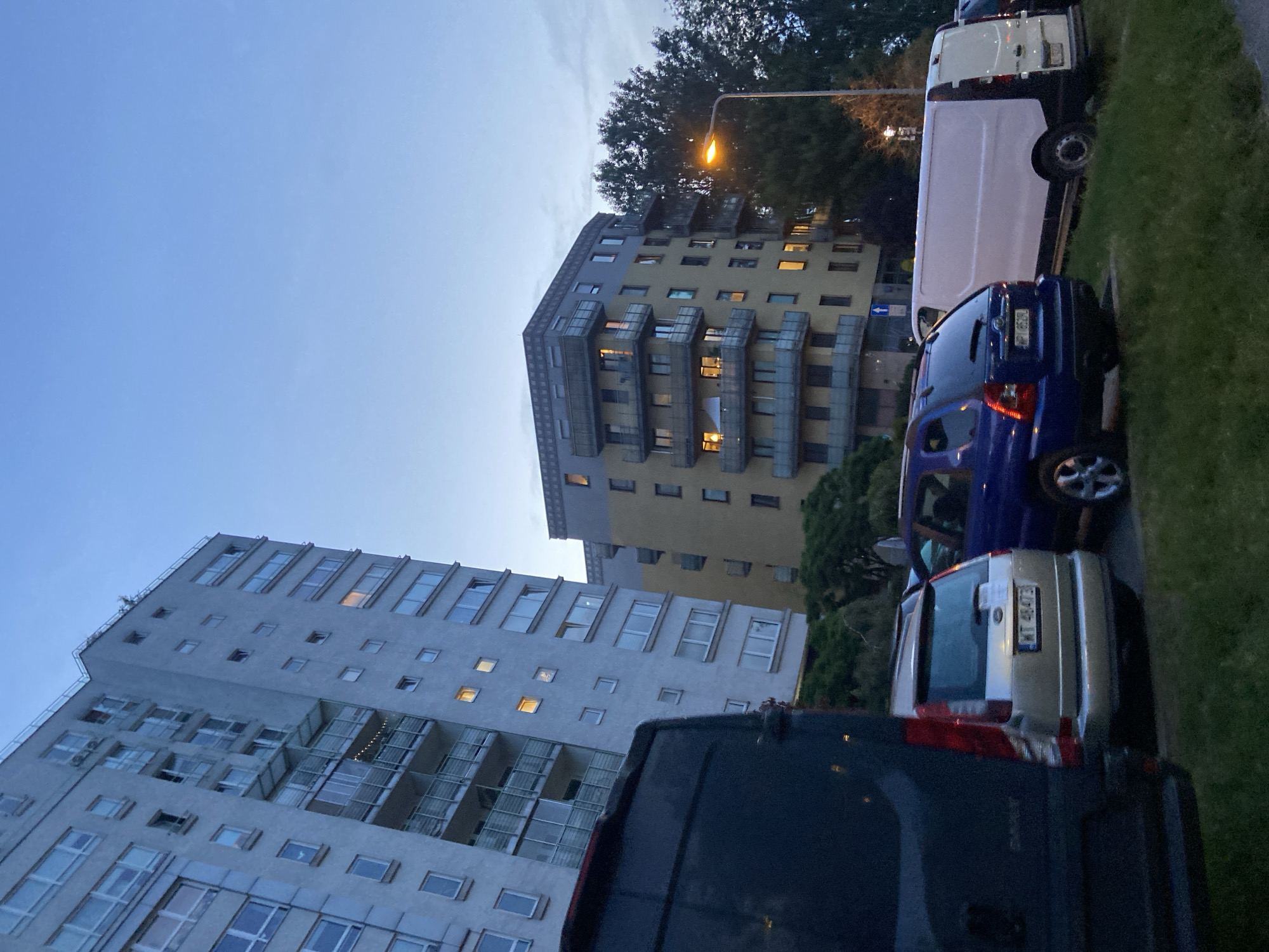 The image depicts an outdoor scene in Warsaw, Poland, likely in the evening as indicated by the twilight sky and illuminated windows in the buildings. In the foreground, the rear of a dark-colored van is visible on the left. To its right, a silver SUV is parked, with its license plate reading "WT 48473". Next to the SUV, a blue hatchback is parked. Further to the right, a white van is parked.
Behind the vehicles, two apartment buildings dominate the scene. The taller building on the left is light grey with numerous windows and balconies. Some windows are lit, suggesting occupancy. The shorter building on the right is yellow with balconies and windows, some of which are also illuminated. A street lamp with a visible orange light is positioned to the right of the yellow building, casting light on the surrounding trees and grass. The environment appears to be a residential area with parked vehicles and trees.