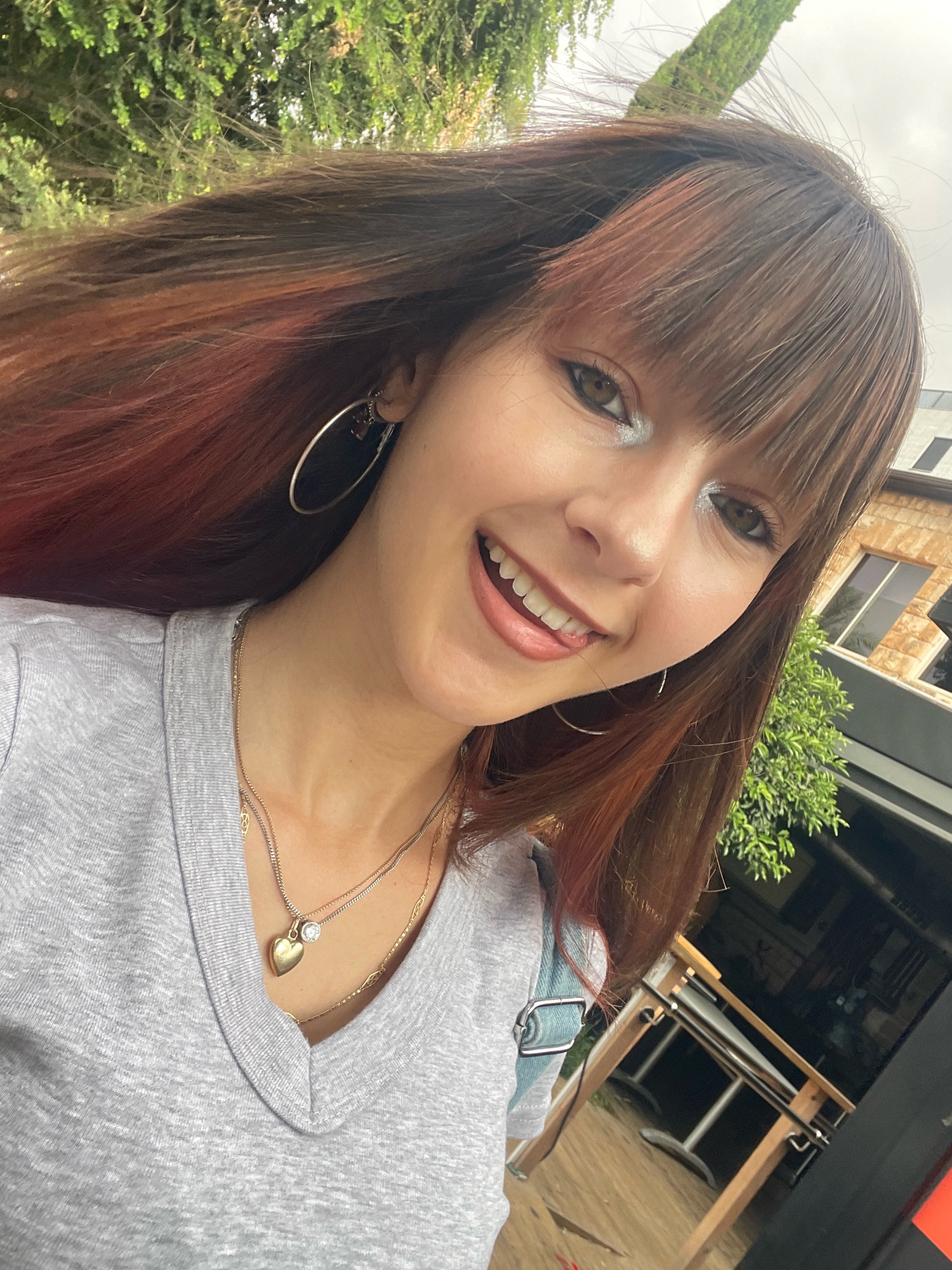 A young woman with long, brown hair with reddish highlights and bangs takes a selfie outdoors. She is wearing a gray v-neck t-shirt and several necklaces, one of which has a heart-shaped pendant. She is also wearing large hoop earrings. Her makeup includes light blue eyeshadow and winged eyeliner. She is smiling with her tongue slightly out, and her left eye is partially closed. Behind her, there is greenery from trees and a building with windows. The scene appears to be in an outdoor urban setting, possibly Raanana, Israel, given the context.