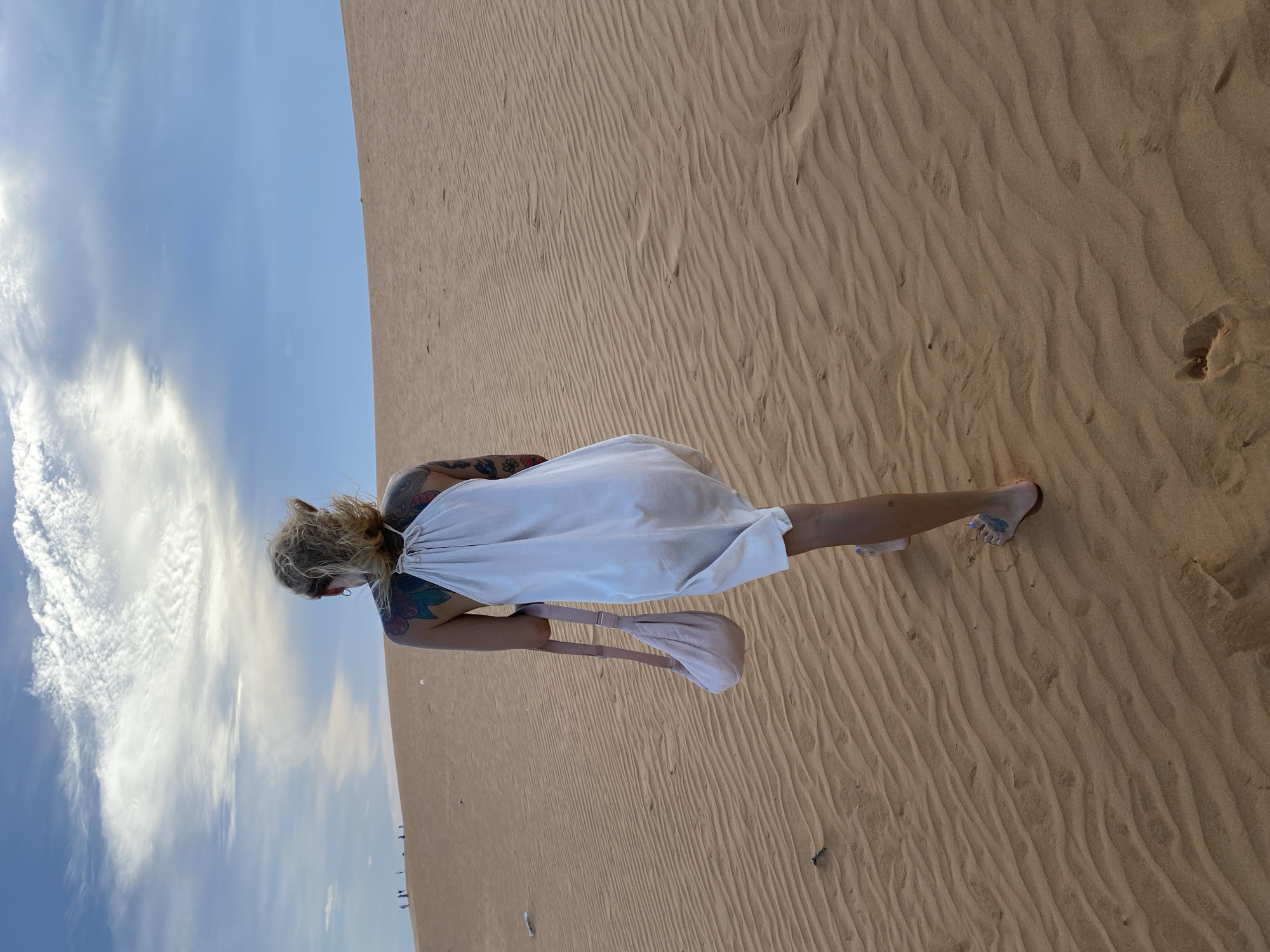 The image displays a person standing on vast sand dunes under a clear blue sky with scattered white clouds, characteristic of the Mui Ne region in Vietnam. The primary subject is a woman, viewed from a slightly high angle, with her back partially turned as she appears to look downwards. She is wearing a simple, flowing light-colored (likely white or off-white) dress and has prominent, colorful tattoos visible on her arms and shoulders. A light pink or beige bag is carried on her left arm. Her bare feet are visible, adorned with blue nail polish, with one foot partially submerged in the soft sand.
The sand dunes stretch across the scene, exhibiting distinct, wavy ripples and ridges formed by wind, rendered in a warm, light brown color. In the far distance, along the horizon where the sand meets the sky, faint figures or structures can be discerned. The overall impression is a serene, outdoor moment of exploration or contemplation in a desert or coastal dune environment. The lighting suggests it is daytime, with bright but not harsh sunlight. There is no visible text in the image.
