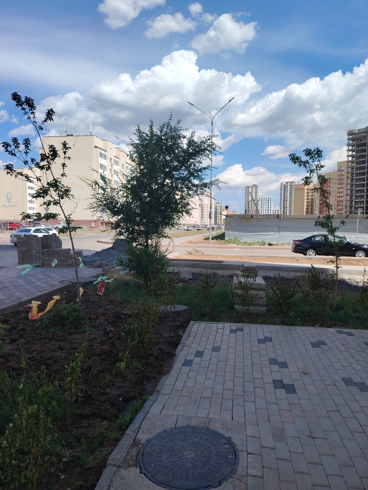 A sidewalk with a manhole cover in the foreground leads to a street lined with new buildings and under construction sites. The sky is bright blue with fluffy white clouds, indicating daytime and fair weather. On the street, a black car is driving and a white car is parked. There are stacks of paving stones and piles of gravel, suggesting ongoing construction or landscaping work. Small trees and bushes are planted along the sidewalk. In the background, several modern apartment buildings, some completed and others still under construction, rise against the sky. A street lamp stands on the left side of the image. The scene appears to be a residential area undergoing development. No people are visible in the frame. No readable text is discernible.