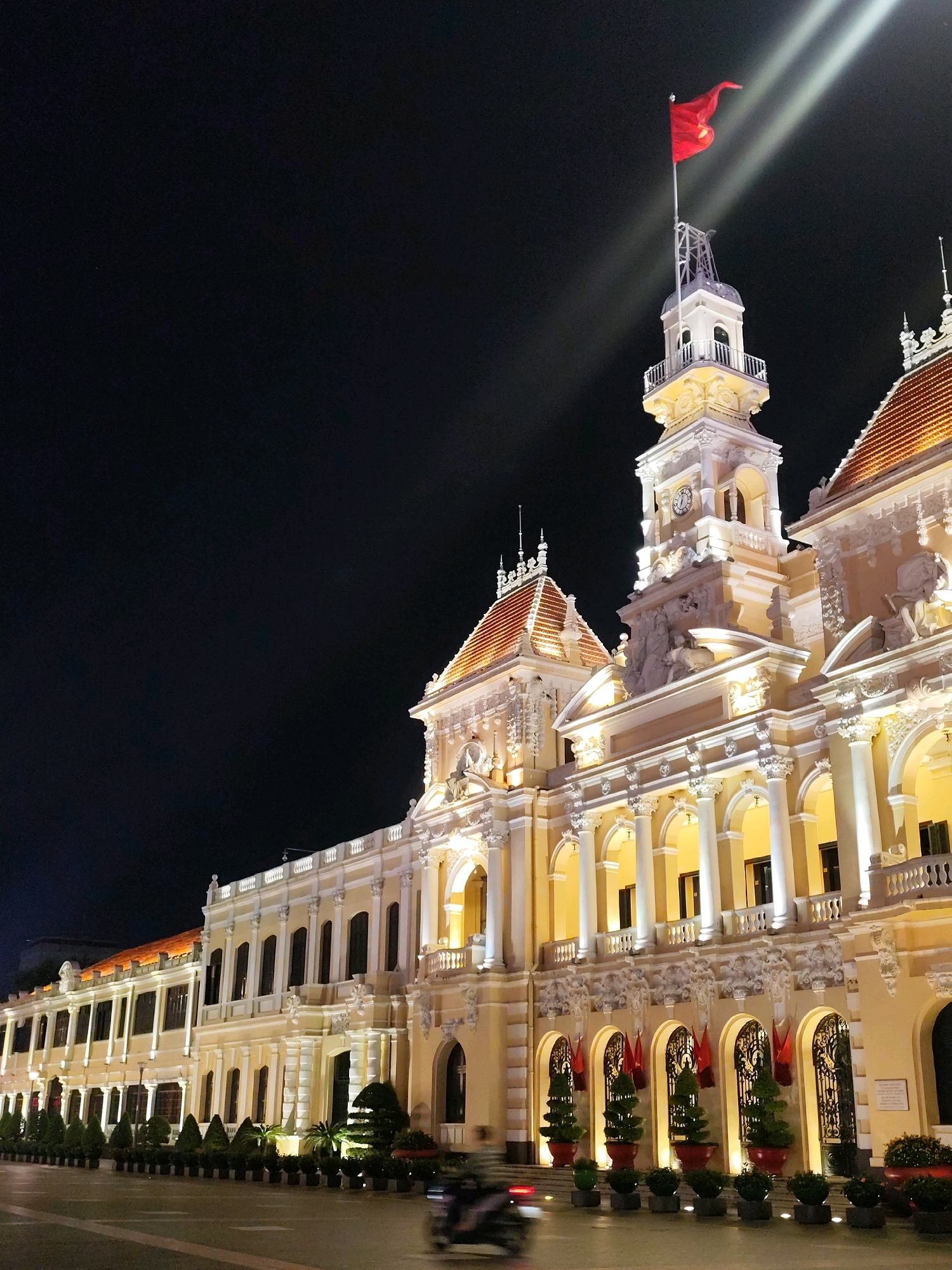 The image shows the Ho Chi Minh City Hall at night. The building is illuminated with warm yellow lights, highlighting its neoclassical architecture. A Vietnamese flag flies from the top of the central tower, illuminated by spotlights. The sky is dark, indicating it is nighttime. In the foreground, a street is visible with a blurred motorcycle in motion. The building features ornate details, including columns, arches, sculptures, and decorative elements. Red flags are displayed along the ground floor arcades, interspersed with potted trees. The overall scene depicts a well-lit, grand building at night in an urban setting.