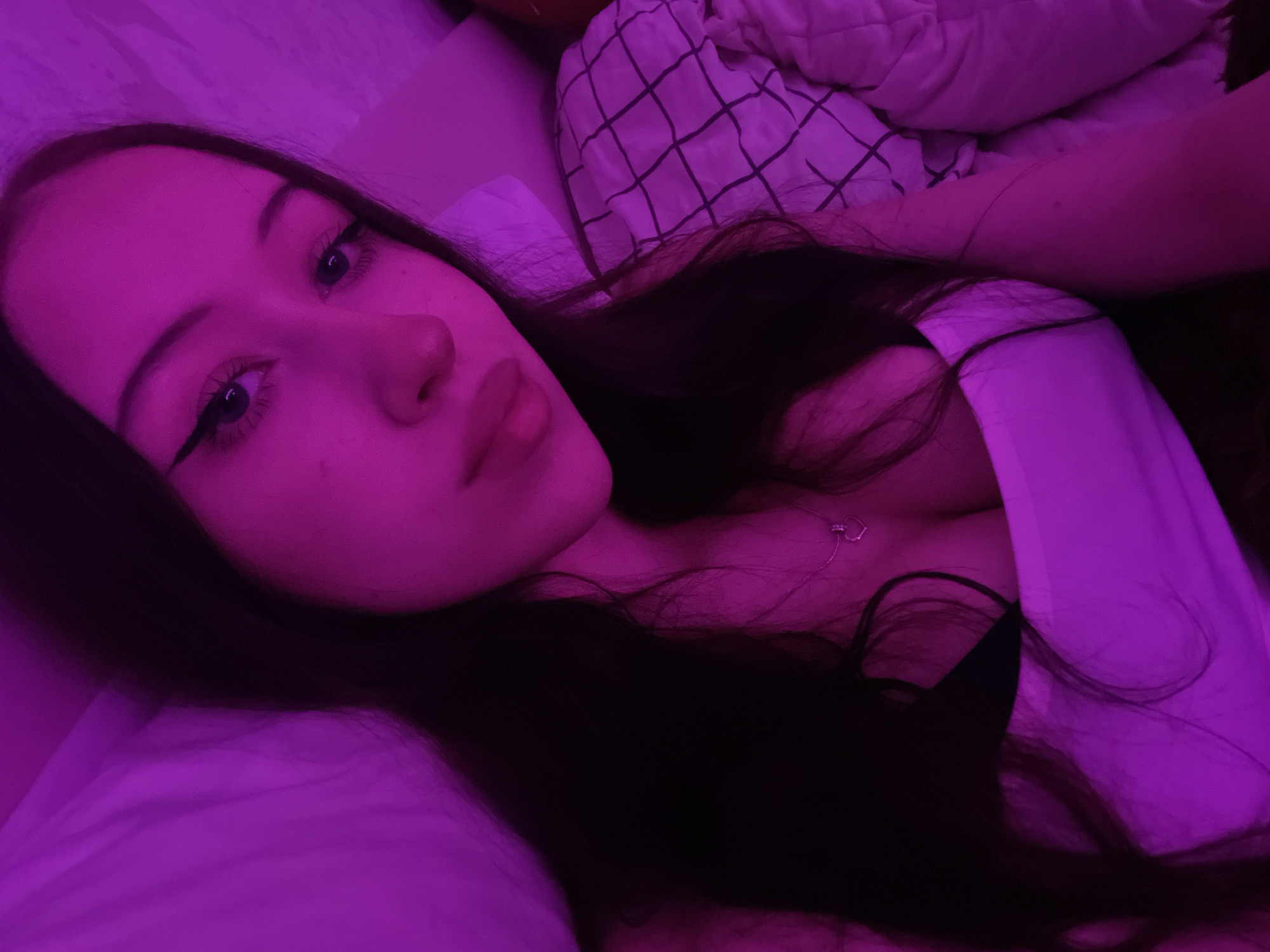 The image shows a young woman with long dark hair, wearing a white tank top and a black bra. She has on winged eyeliner and a silver necklace with a heart pendant. She is lying in bed, partially covered by a blanket with a black checkered pattern. The lighting in the room is a vibrant pink. The image does not contain any information that would allow for its location to be identified as Votkinsk, Russia.