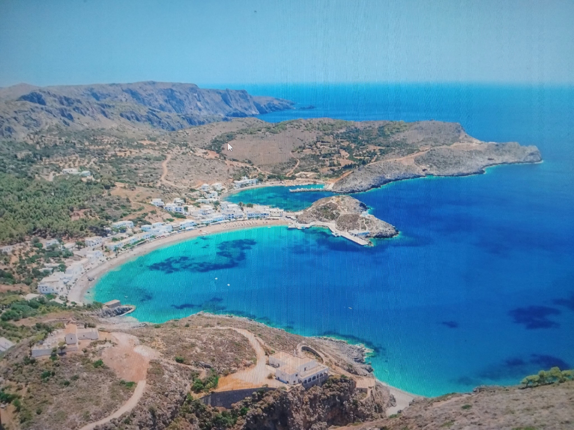 The image shows an aerial view of a coastal landscape. A crescent-shaped bay is lined with white buildings, likely a town or village. A sandy beach borders the turquoise water within the bay. Rocky cliffs and headlands surround the bay, with sparse vegetation covering the land. Mountains are visible in the background. The water transitions to a deeper blue further out from the bay. The location is not in Belene, Bulgaria, but appears to be a coastal area possibly in the Mediterranean region.
