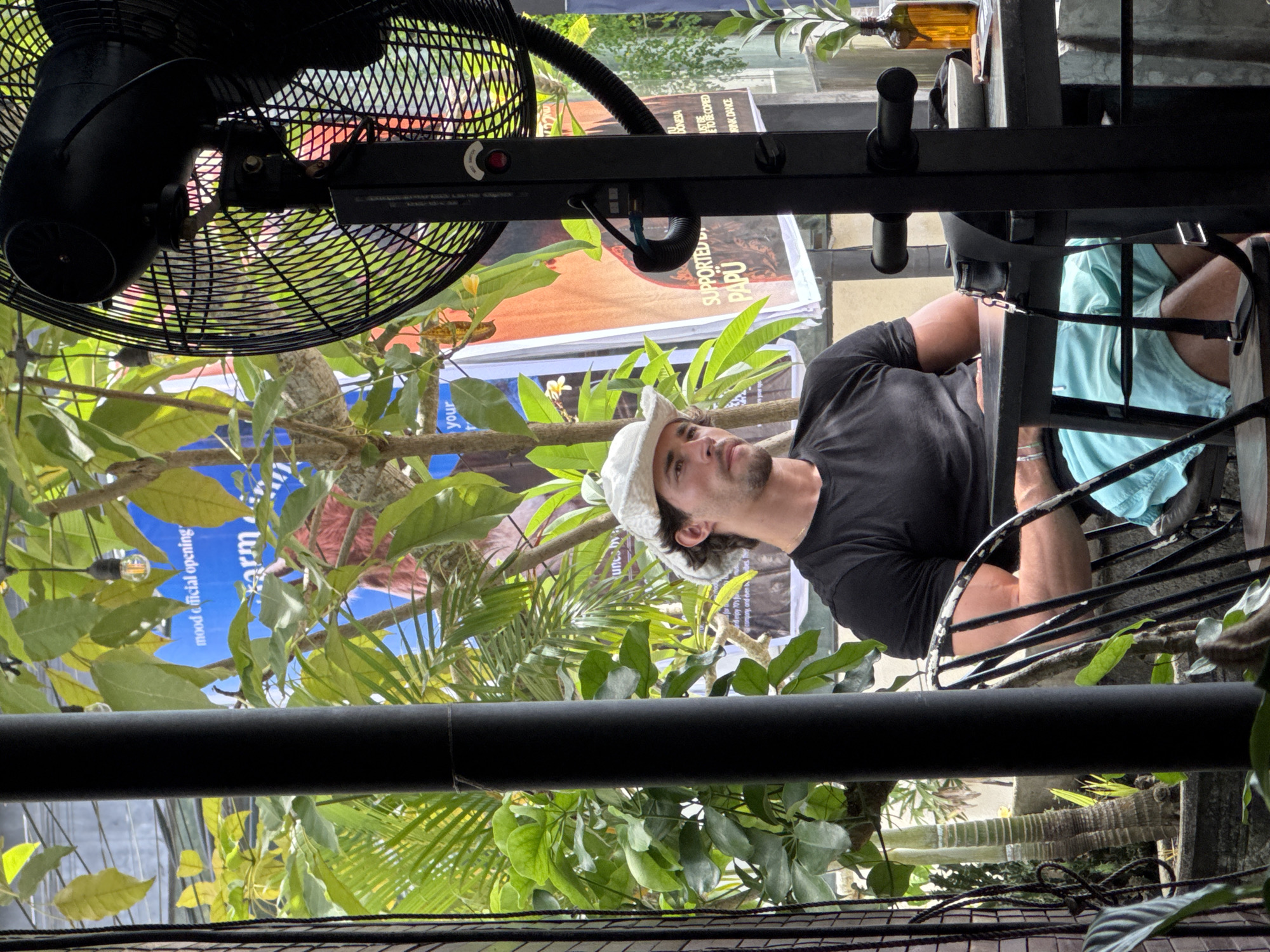 The image depicts a scene in Kuta Selatan, Indonesia. A man wearing a white hat and black shirt sits on a chair outdoors. A person wearing light blue shorts sits next to him. In the background are green trees and posters. A black fan hangs overhead. A black iron railing and table surround the scene.