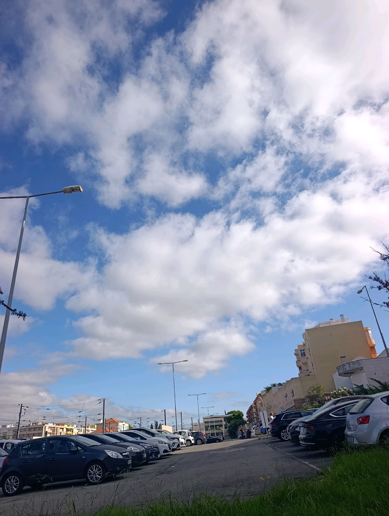 The image shows a street scene in Setúbal, Portugal, featuring a parking lot filled with cars and a building in the background. The sky is blue with scattered white clouds. A street lamp is visible on the left side of the image. The parking lot is lined with several cars, including dark-colored sedans and a lighter-colored hatchback in the foreground. In the background, there is a multi-story building with a light-colored facade. The scene appears to be during the daytime.