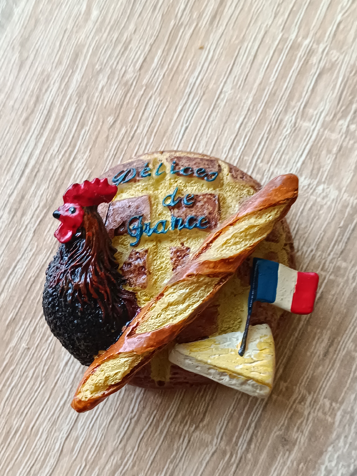 This is a close-up, top-down shot of a souvenir magnet shaped like a French-themed food assortment. The magnet is placed on a light-colored, wood-grained surface, likely a table.
The main subjects of the magnet are a stylized French baguette, a wedge of cheese, a rooster, and a small French flag. The baguette is depicted with a golden-brown crust, lying diagonally across the scene. The cheese appears to be a light yellow triangular wedge with a textured rind. The rooster, a common symbol of France, is positioned on the left, rendered in dark brown and black with a vibrant red comb and wattle. A small French flag, with vertical stripes of blue, white, and red, is planted in the cheese.
The background of the magnet is a circular shape with a brick-like texture and yellow highlights, resembling a loaf of bread or perhaps a tart. Blue, cursive text is inscribed on this background. The visible text reads "délices de France".
There is no discernible activity or event taking place, as the subjects are static. The lighting suggests an indoor setting, possibly during daytime, with no specific weather cues. The location context of Tebras, Latvia, is provided, but there are no direct visual cues within the image itself to confirm this location. The overall impression is a decorative item representing French culinary and cultural symbols.